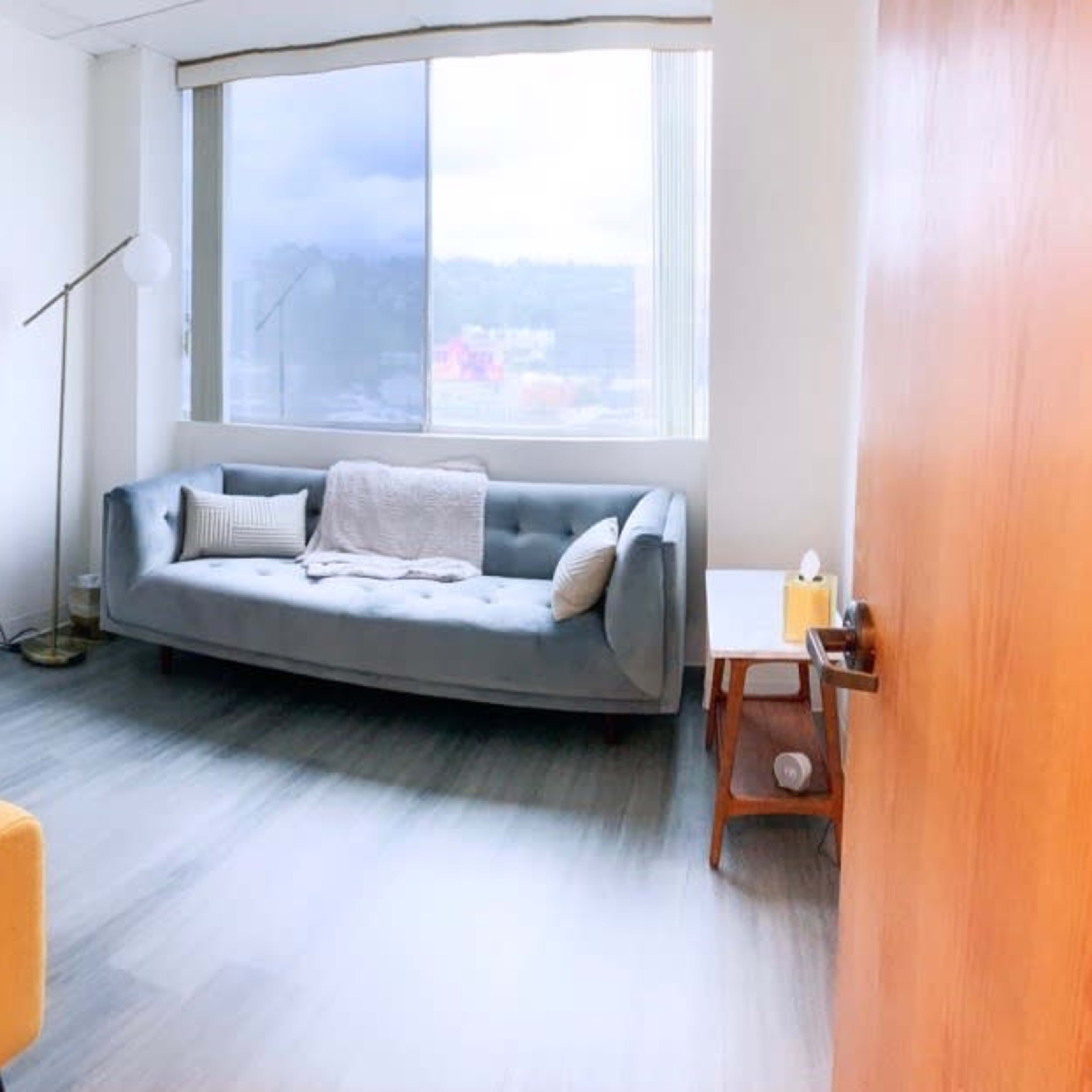 A light-colored room with a blue sofa, a yellow armchair, a floor lamp, and a small side table near a large window.