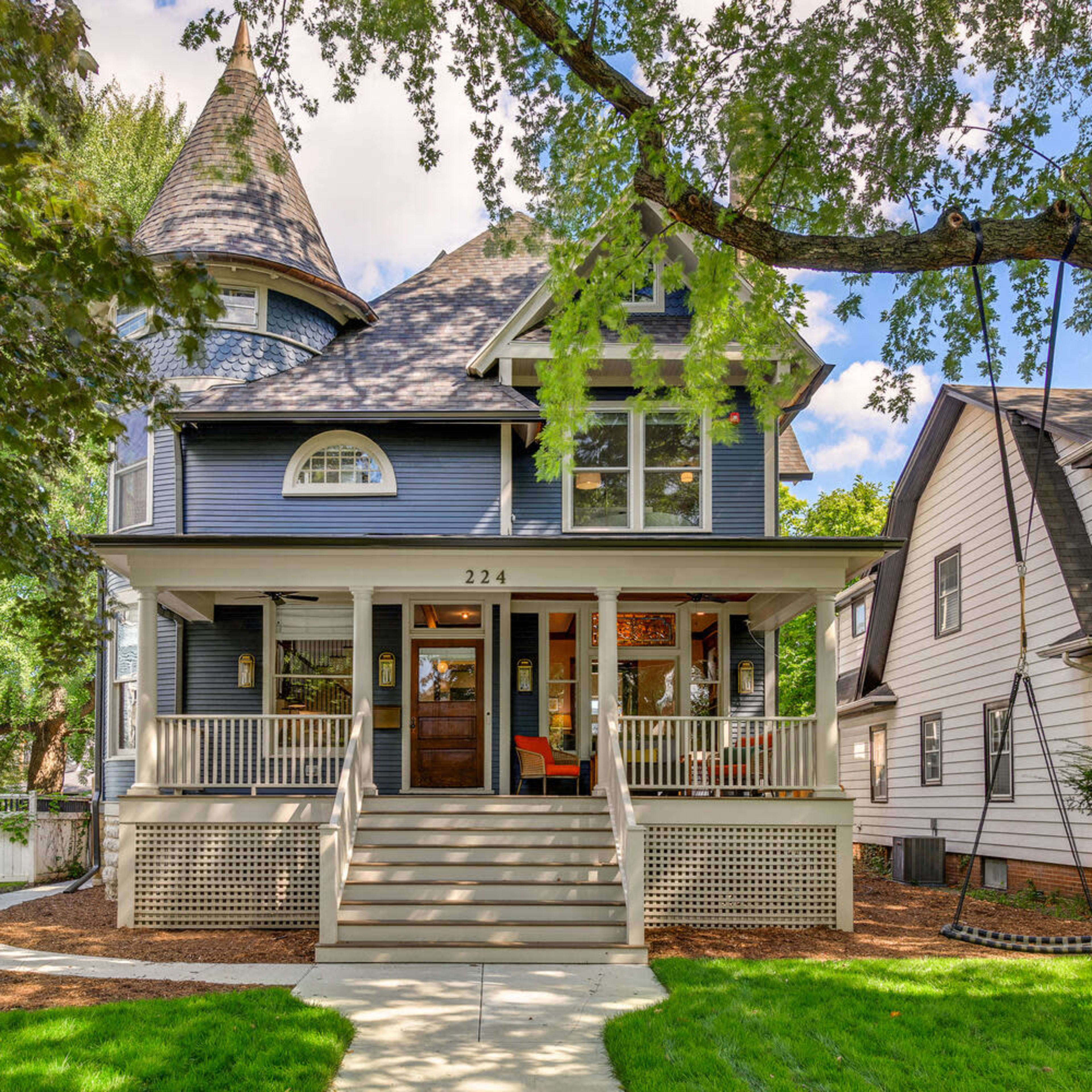 A large, multi-story blue house with a turret and a welcoming front porch is surrounded by trees and lawns.