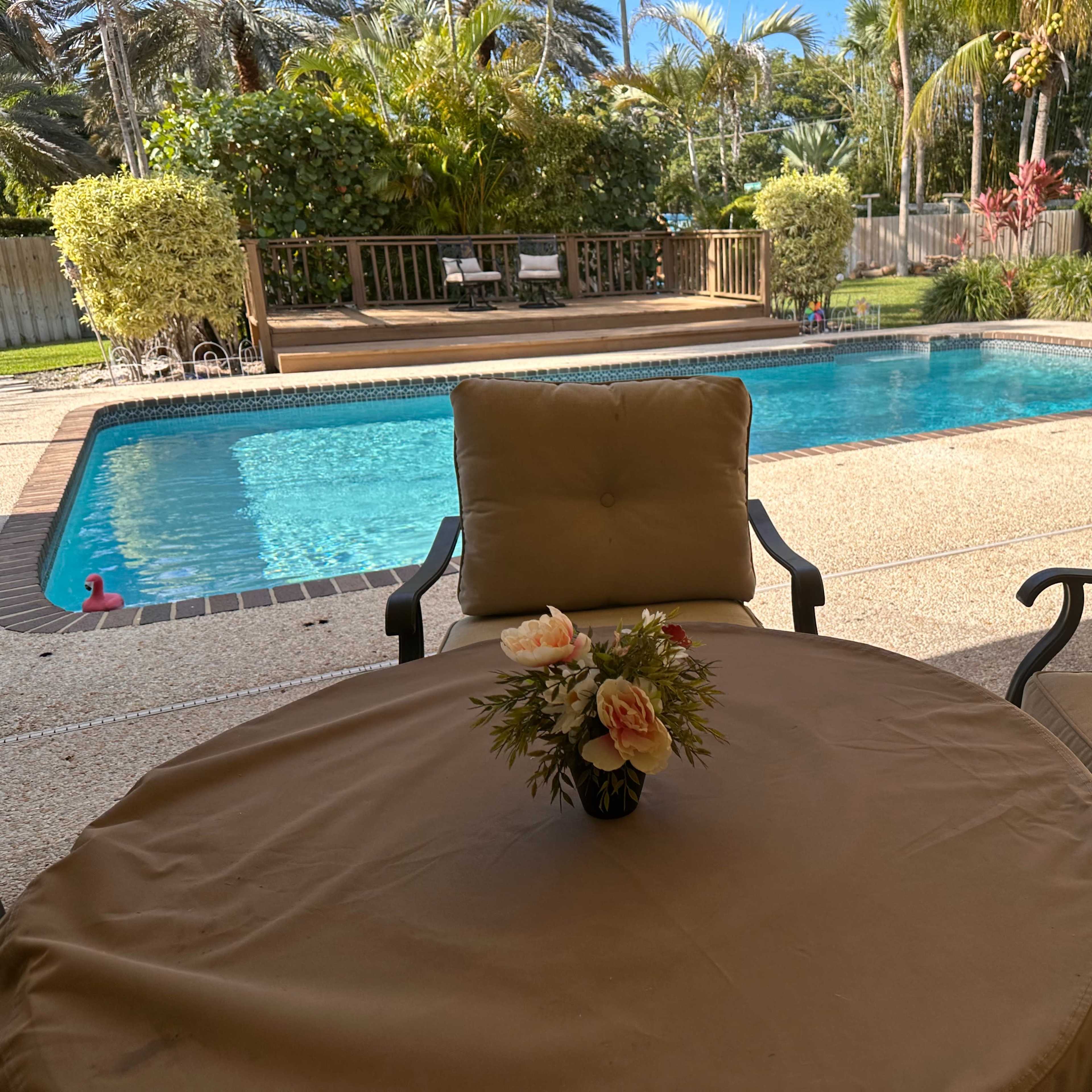 A view of a swimming pool surrounded by a landscaped yard, taken from a seated area with a small table and a flower arrangement.