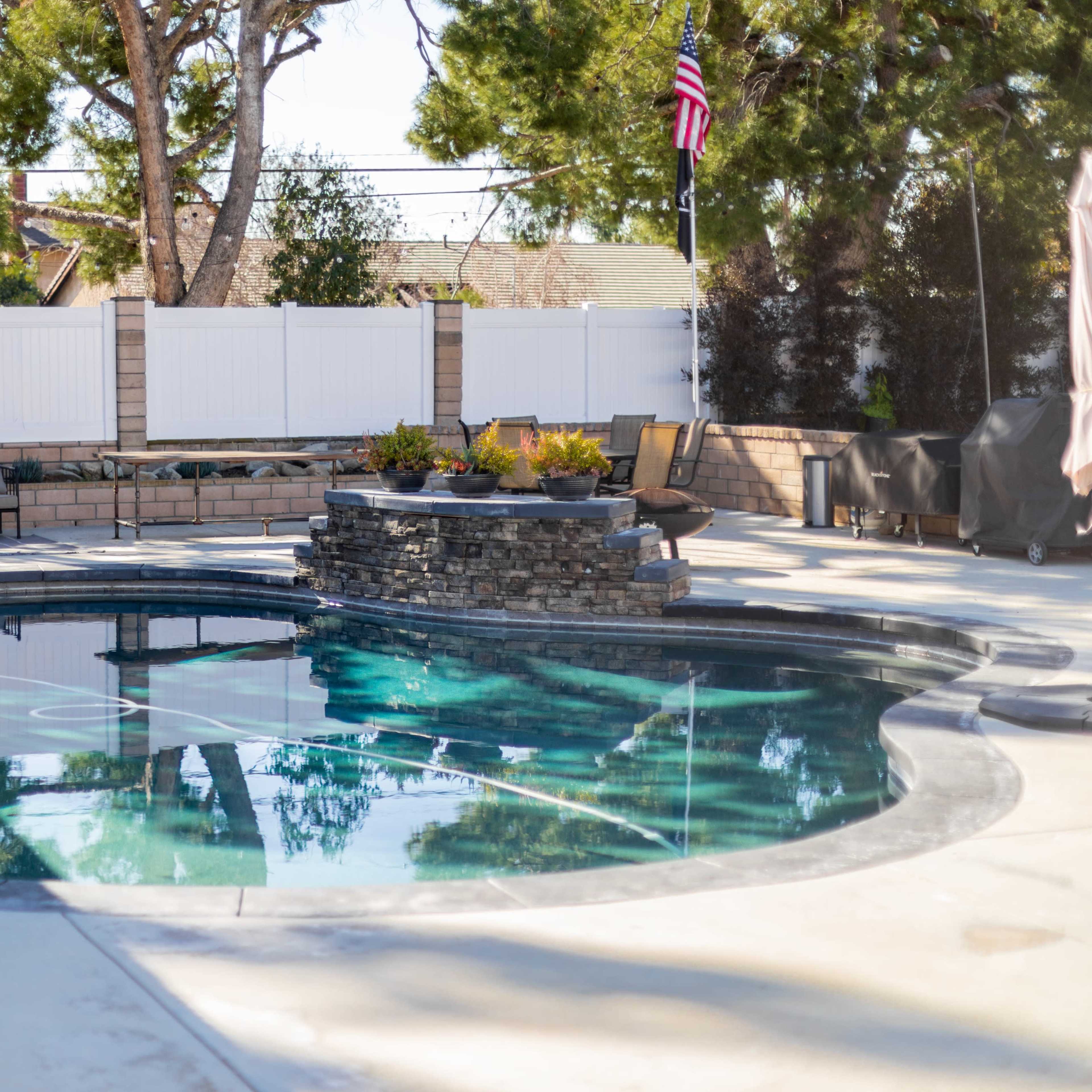 A backyard featuring a Mickey Mouse-shaped swimming pool with a stone fountain, surrounded by lounge furniture and a white fenced area.