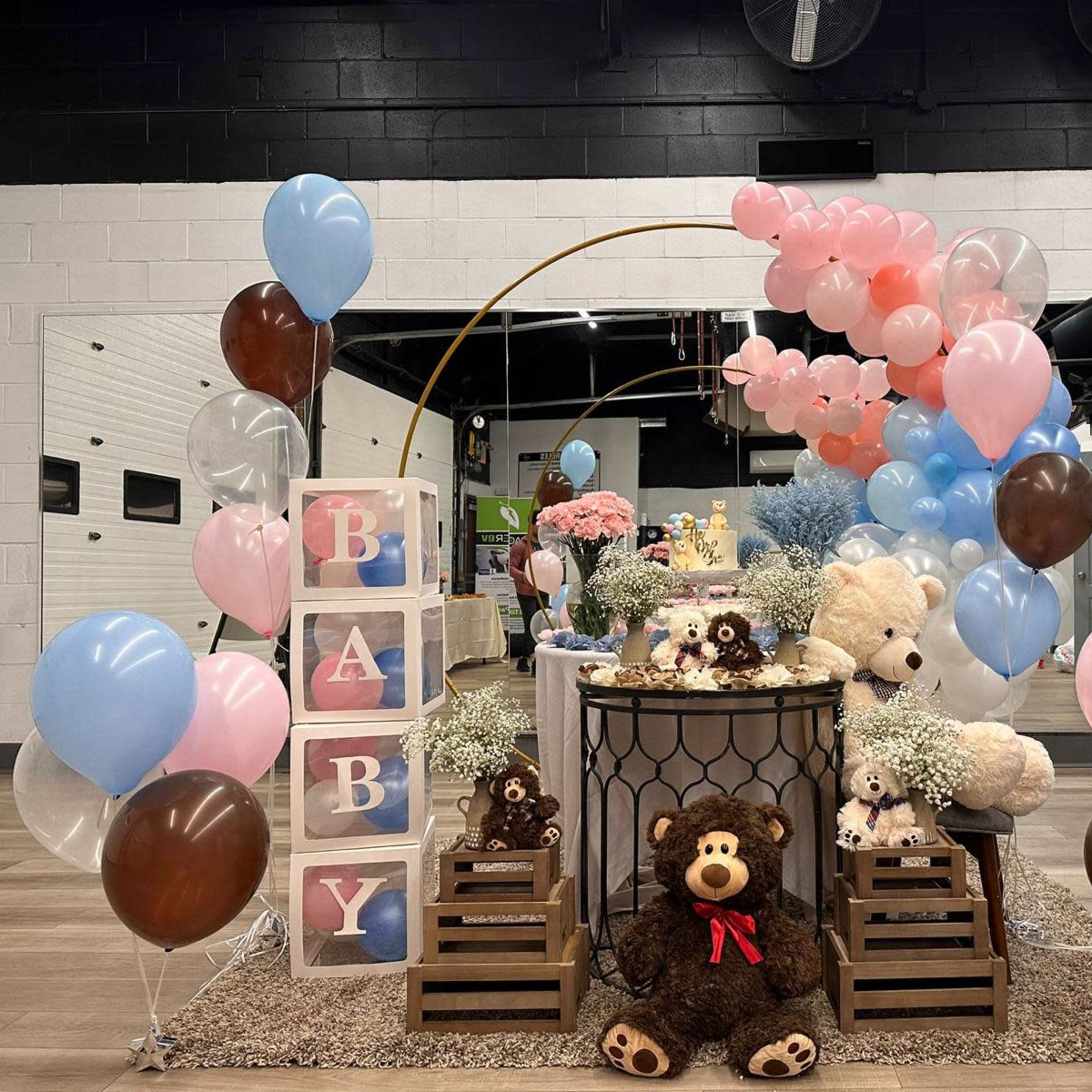 A decorative setup for a baby celebration featuring pastel balloons, stuffed animals, and a dessert table with a cake surrounded by floral arrangements.