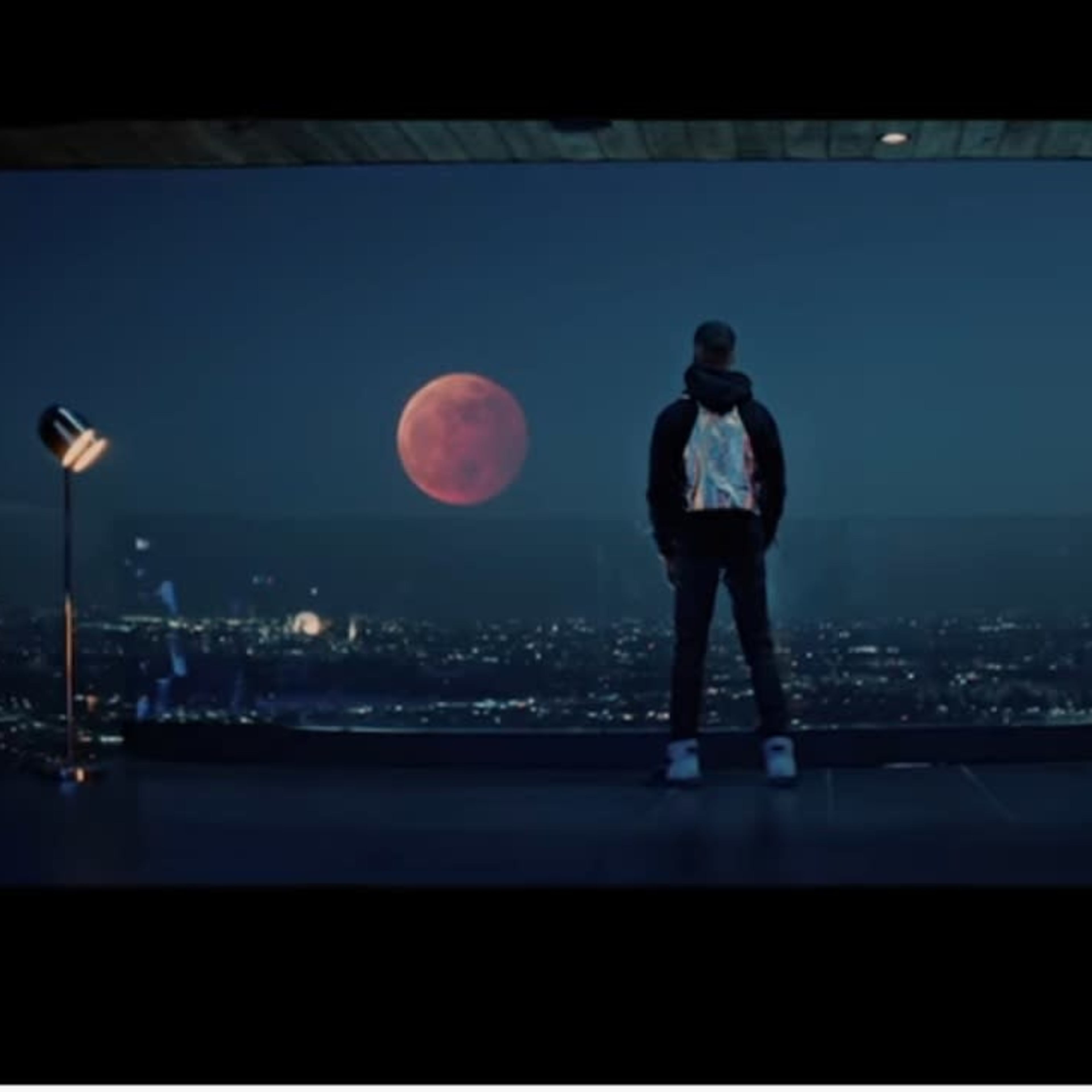 A person stands on a balcony overlooking a city skyline at night, with a large red moon illuminating the sky.