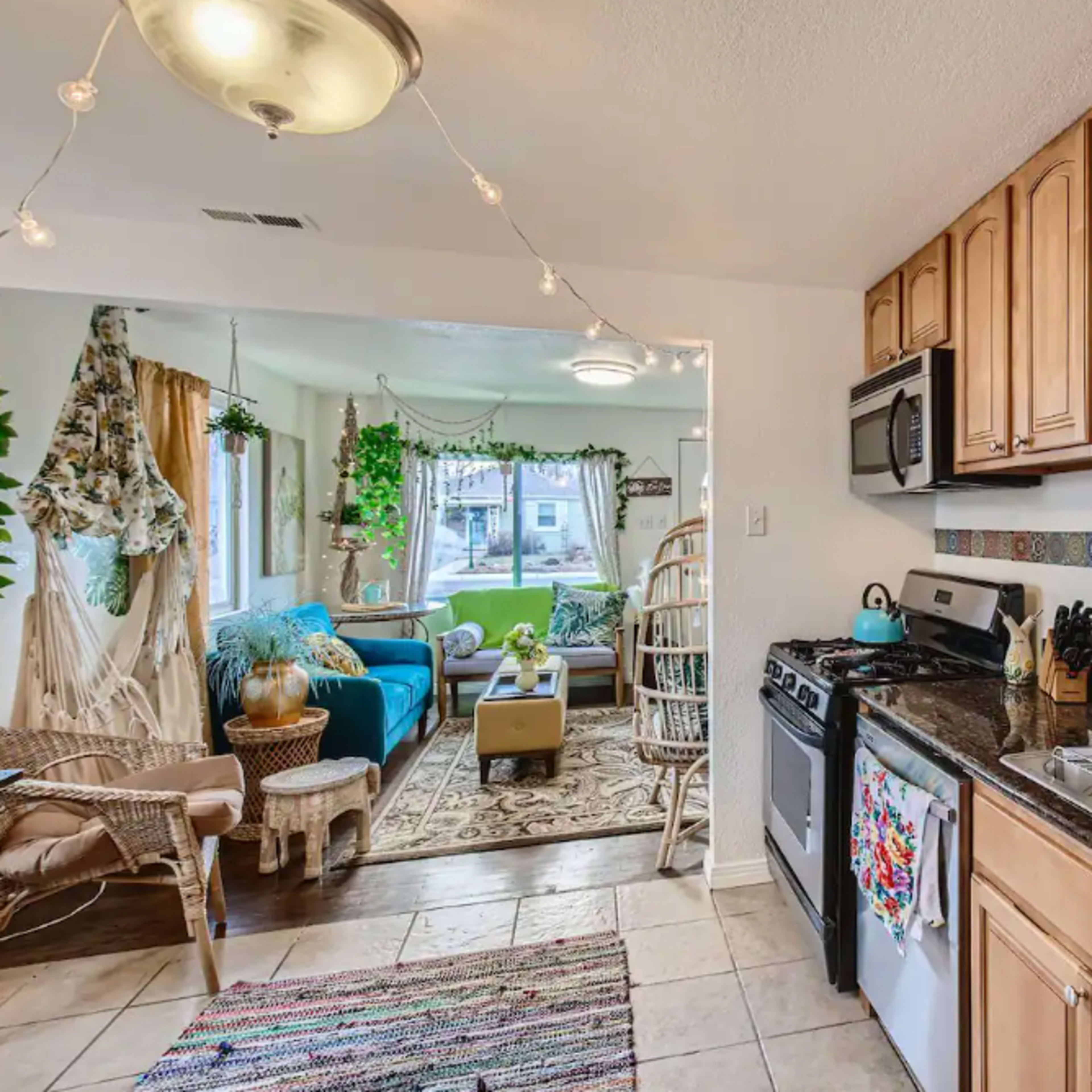 A small kitchen with wooden cabinets, a stainless steel stove, and a black countertop opens into a living area filled with plants and comfortable seating.