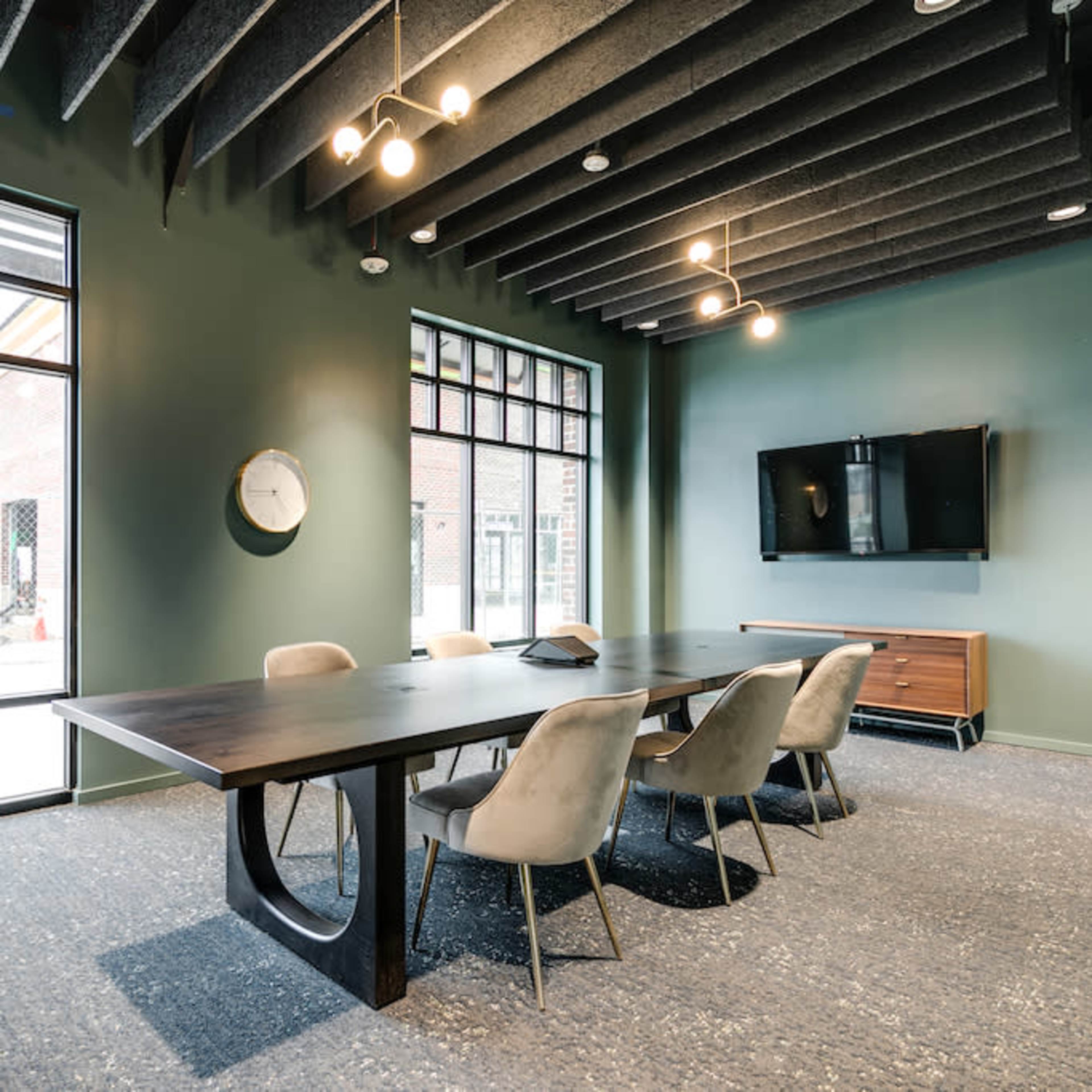 A conference room features a large wooden table surrounded by upholstered chairs, with a wall-mounted television and large windows overlooking a construction site.