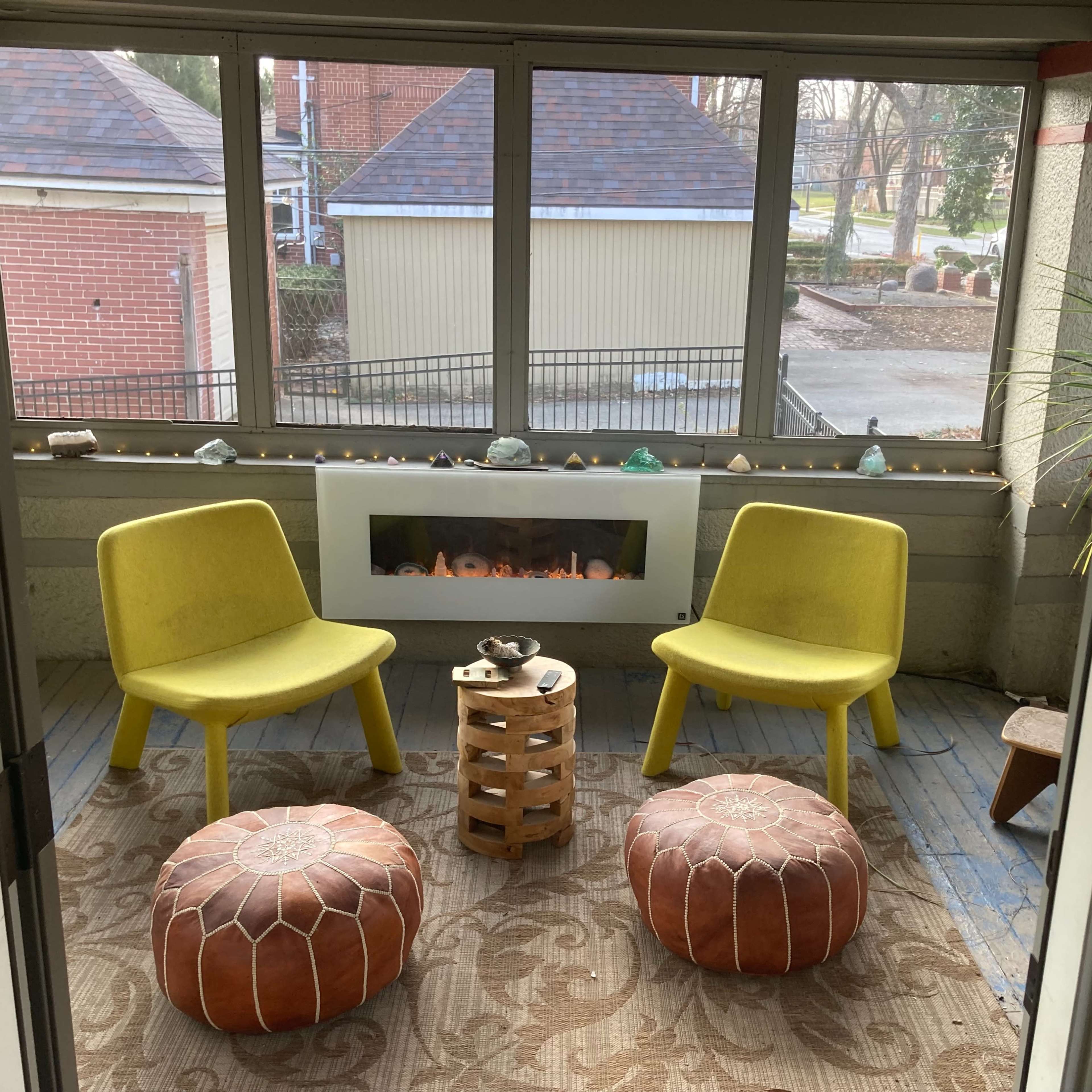 A sunroom with two yellow chairs, a wooden stool, and two decorative poufs arranged around a modern electric fireplace.