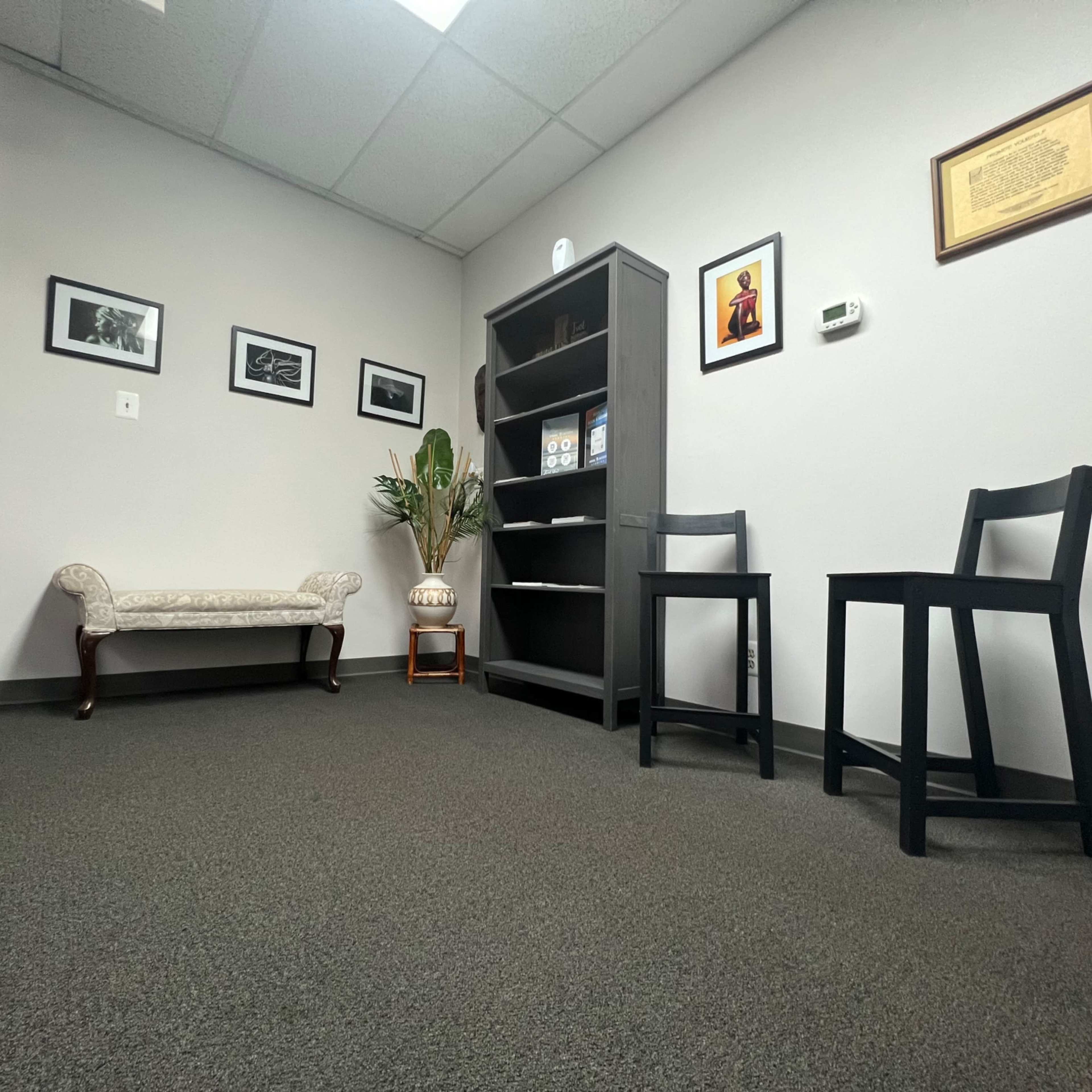 A small, minimalist office space with a decorative chair, a bookshelf, and framed pictures on the walls.