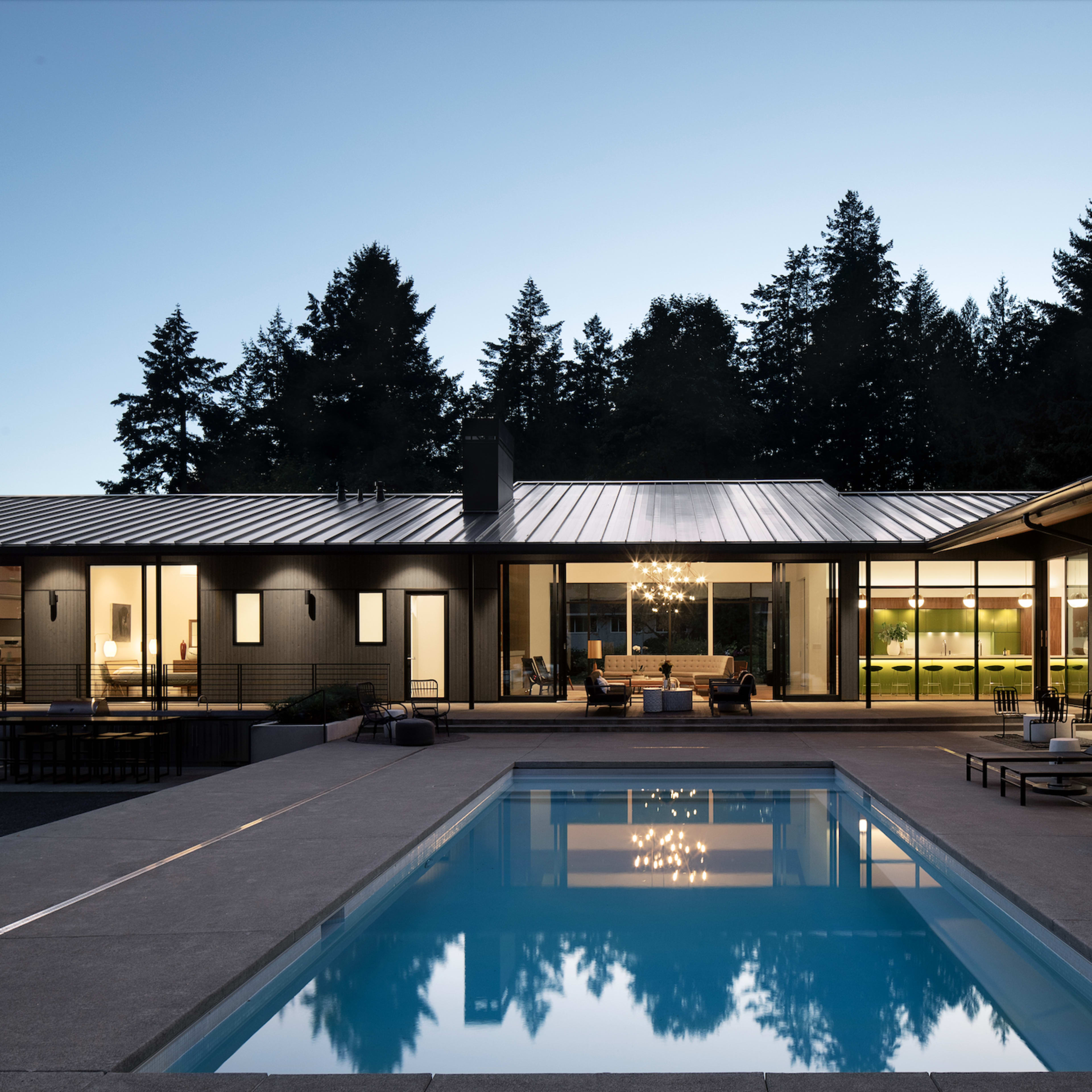 A modern house with large windows surrounds a crystal-clear swimming pool, set against a backdrop of tall trees at twilight.