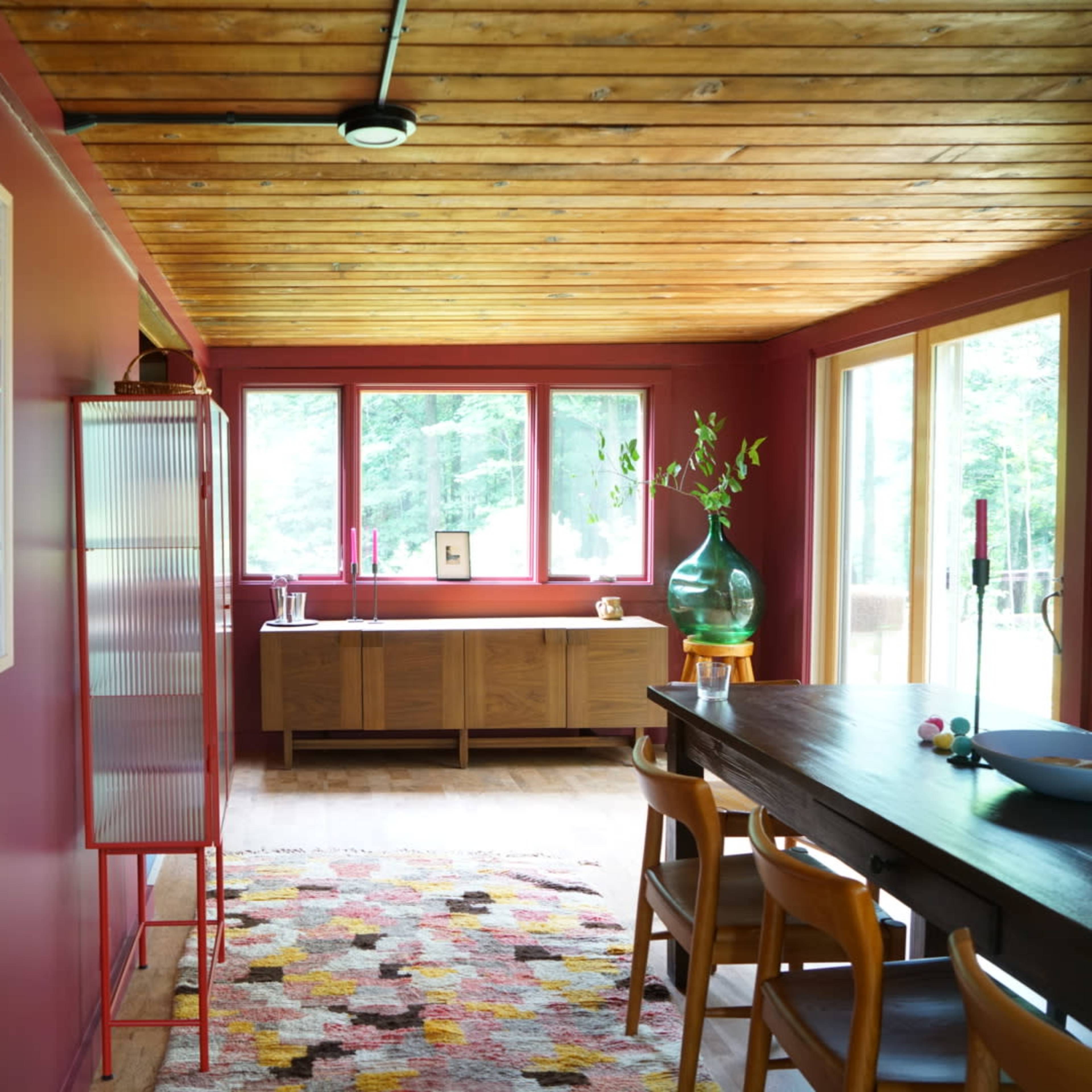 A room with red walls, wooden ceiling, a dining table, and large windows allowing natural light to enter.