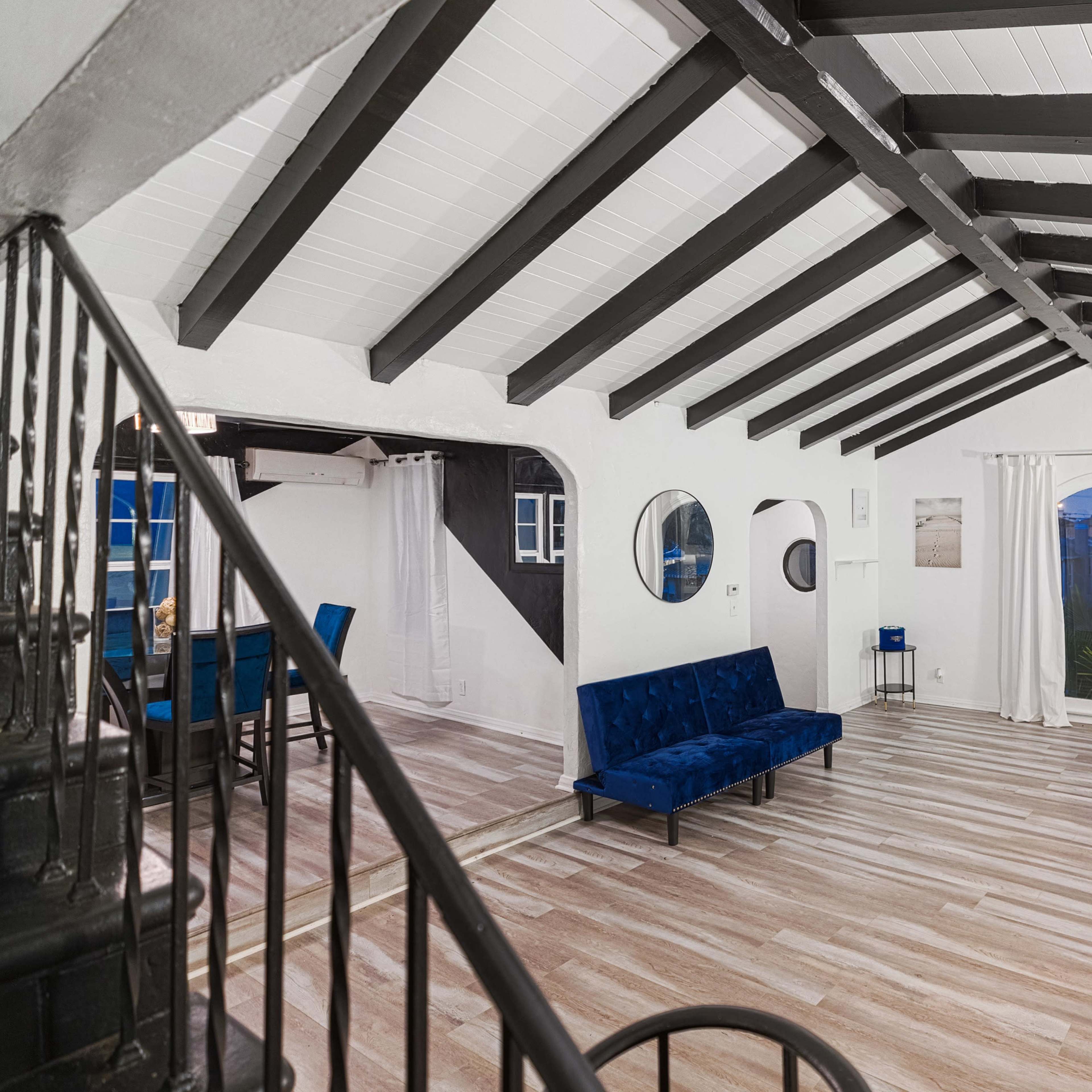 The room features a spacious layout with dark wooden beams, light-colored walls, and blue seating arrangements.