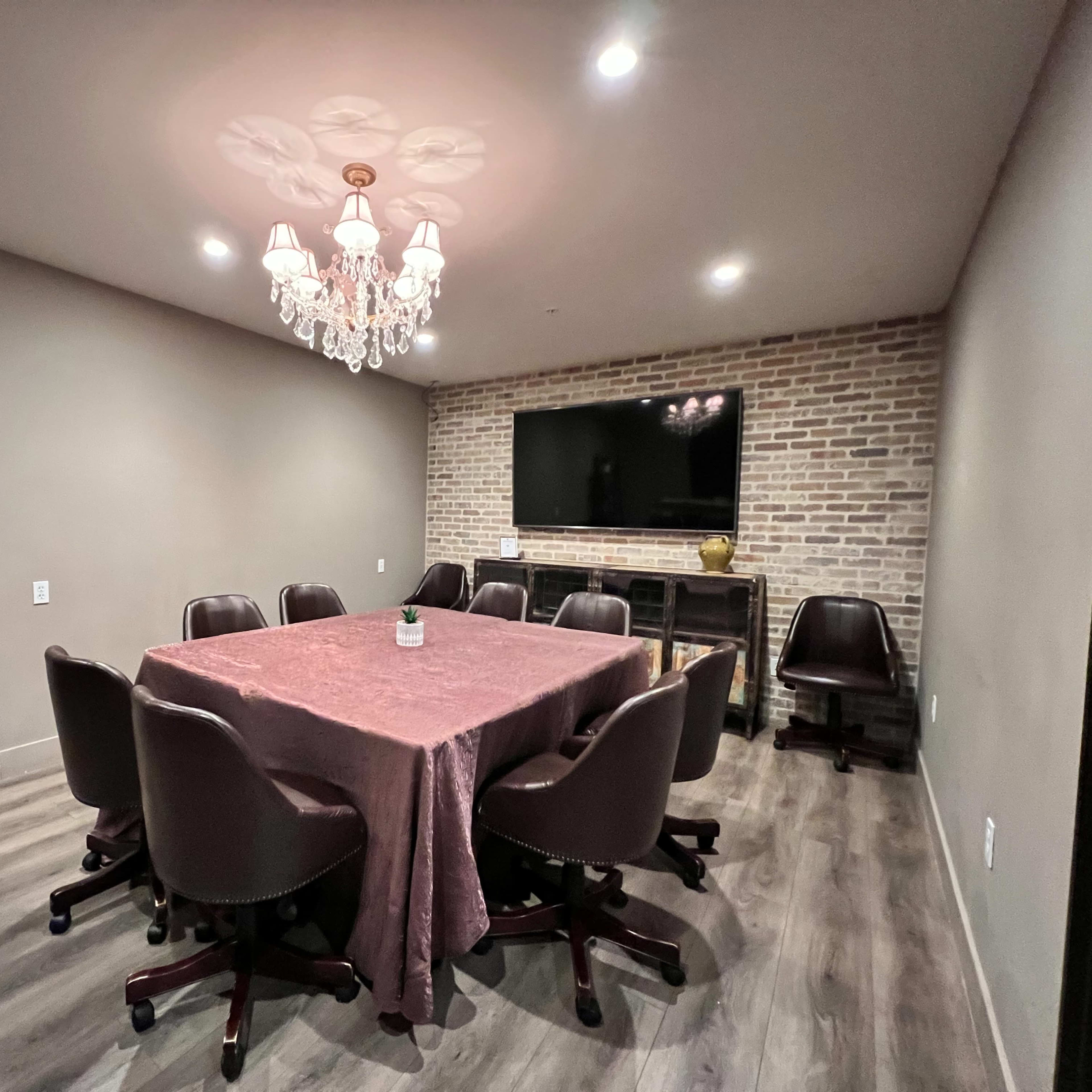 A conference room with a large table surrounded by eight brown chairs, a chandelier above, and a brick wall with a television mounted on it.
