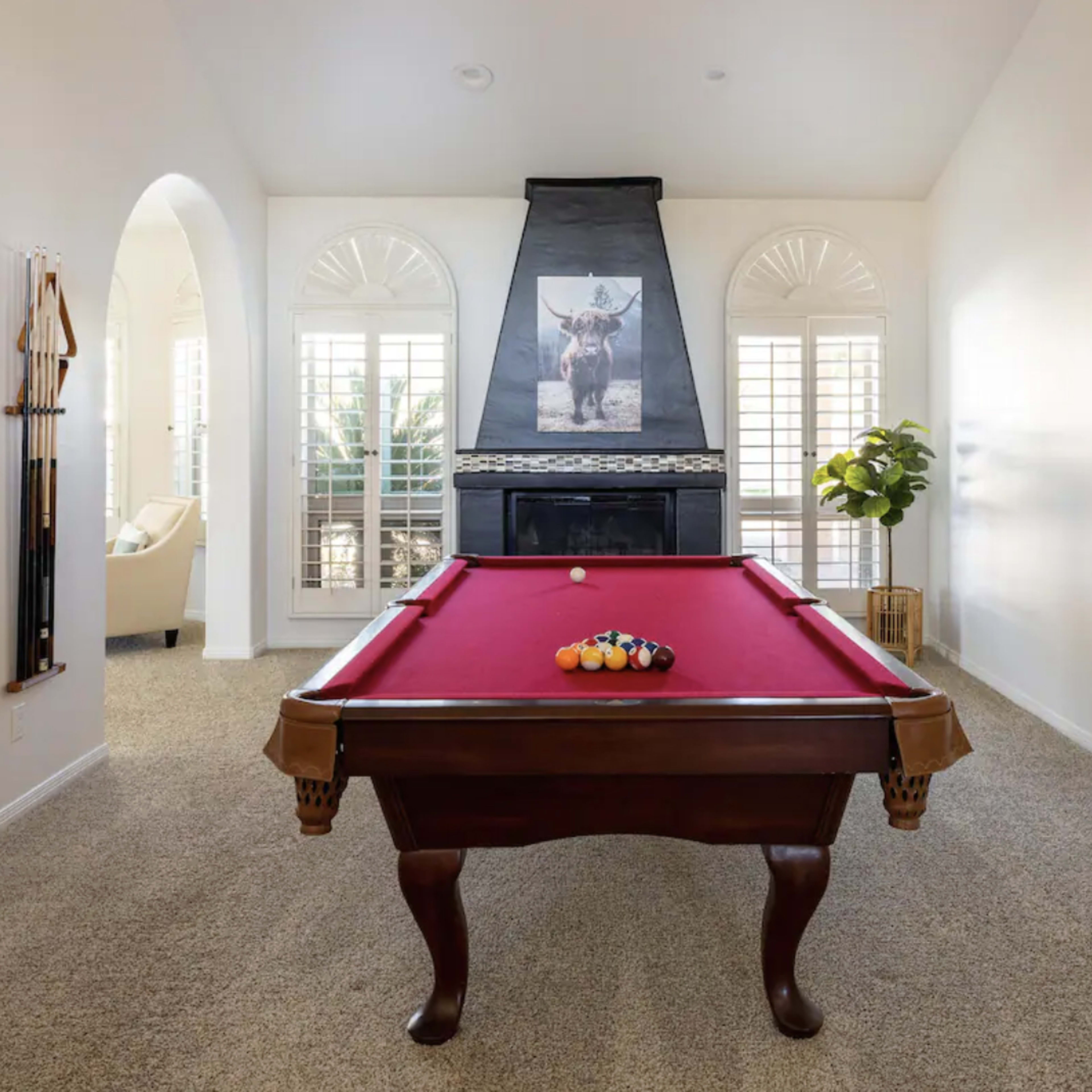 A billiard table with a red felt surface, set in a room with large windows, a decorative fireplace, and a piece of wall art above the mantel.