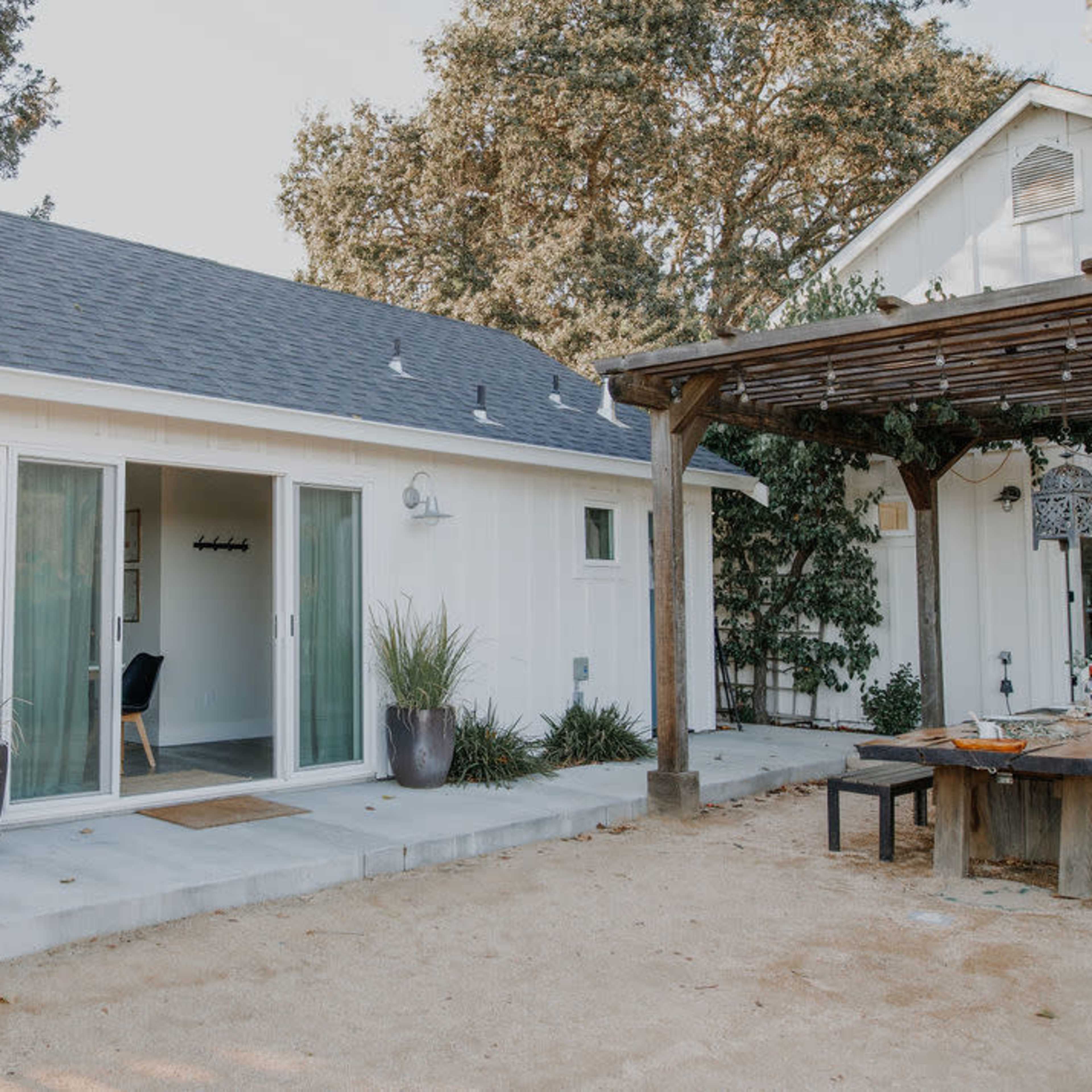 A modern, white cottage with large glass doors opens onto a patio area, surrounded by greenery and a shaded pergola.