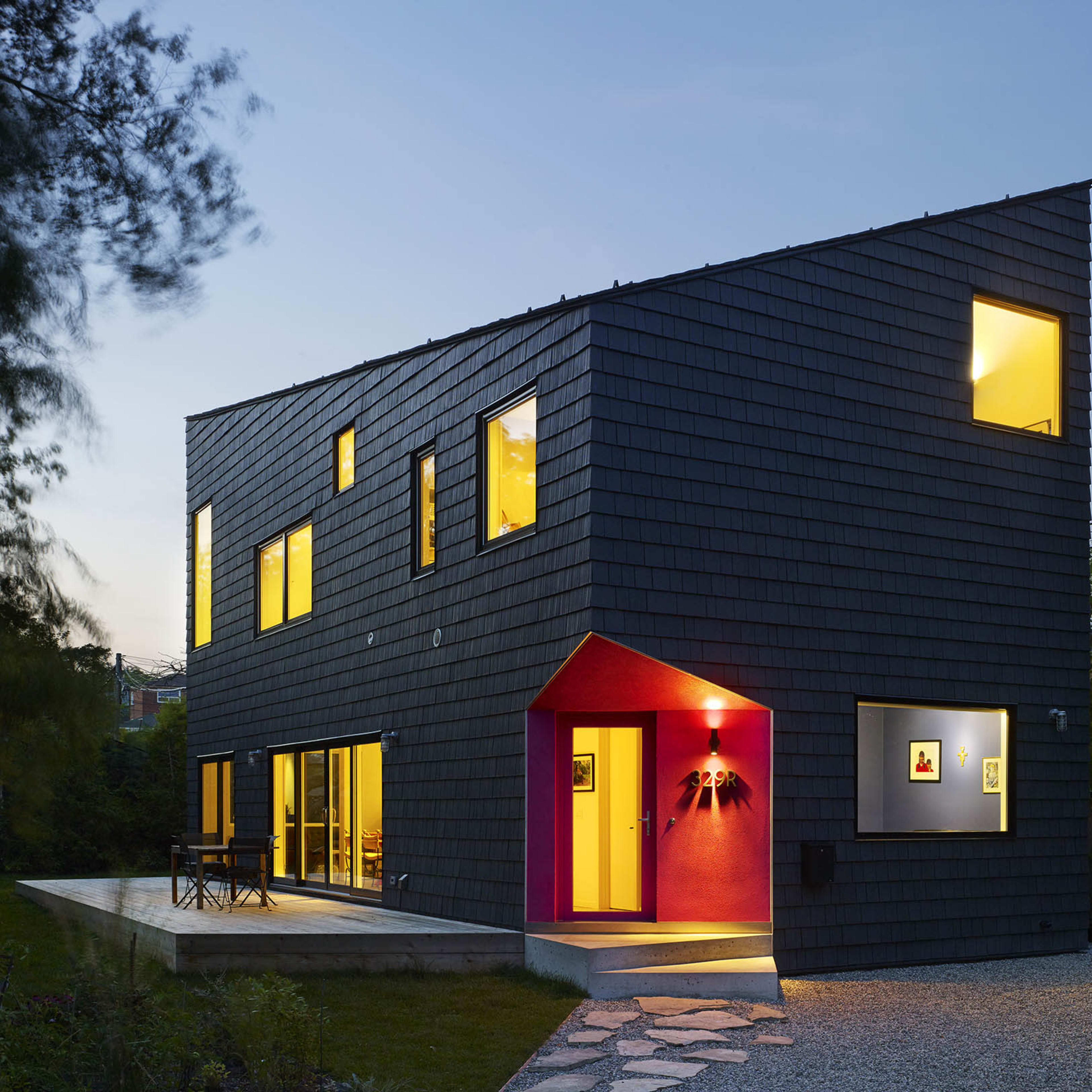 A modern two-story house with a dark shingle exterior and a prominently colored entryway, situated in a landscaped yard at dusk.