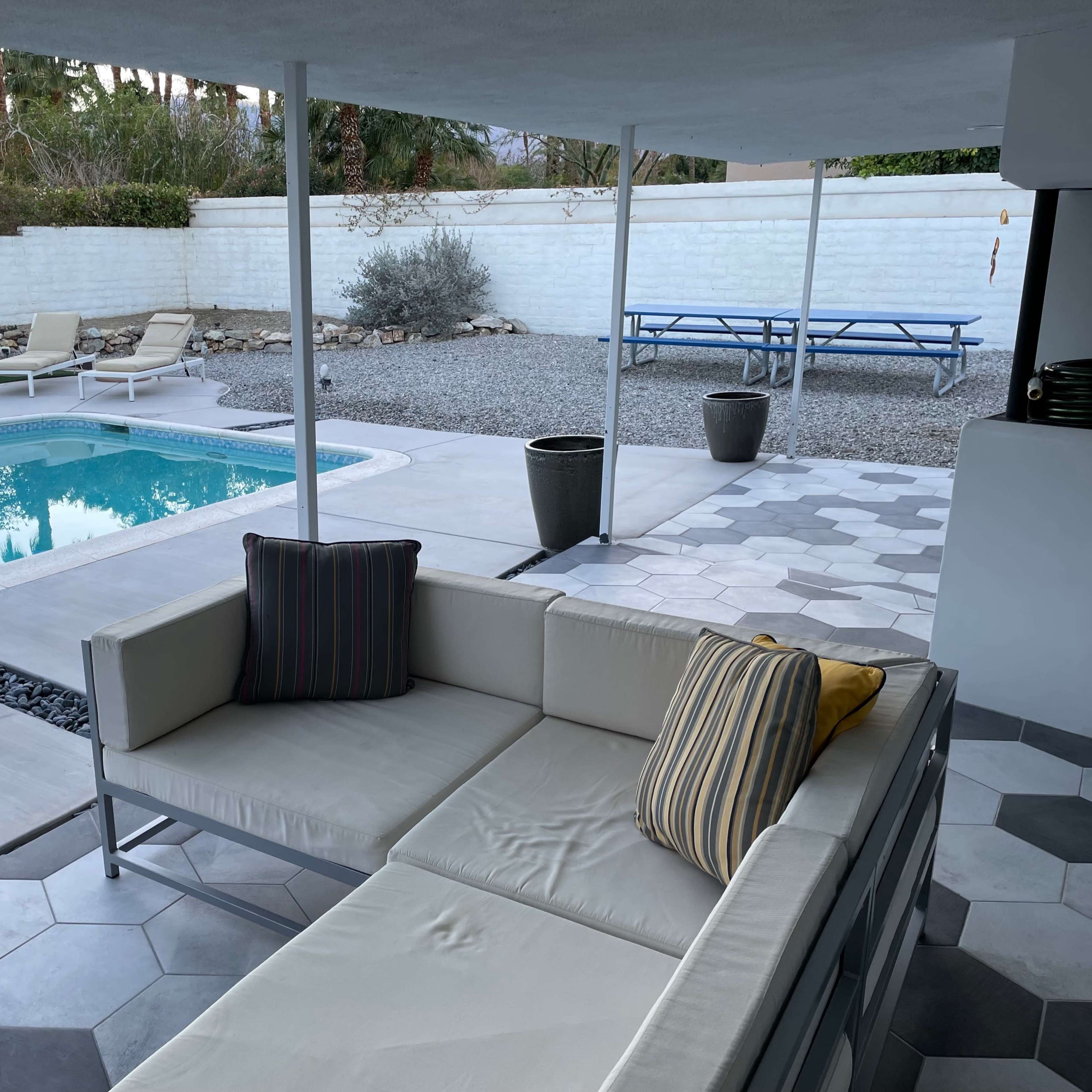 A modern outdoor lounge area beside a swimming pool, featuring a sectional sofa, sun loungers, and a gravel landscape.