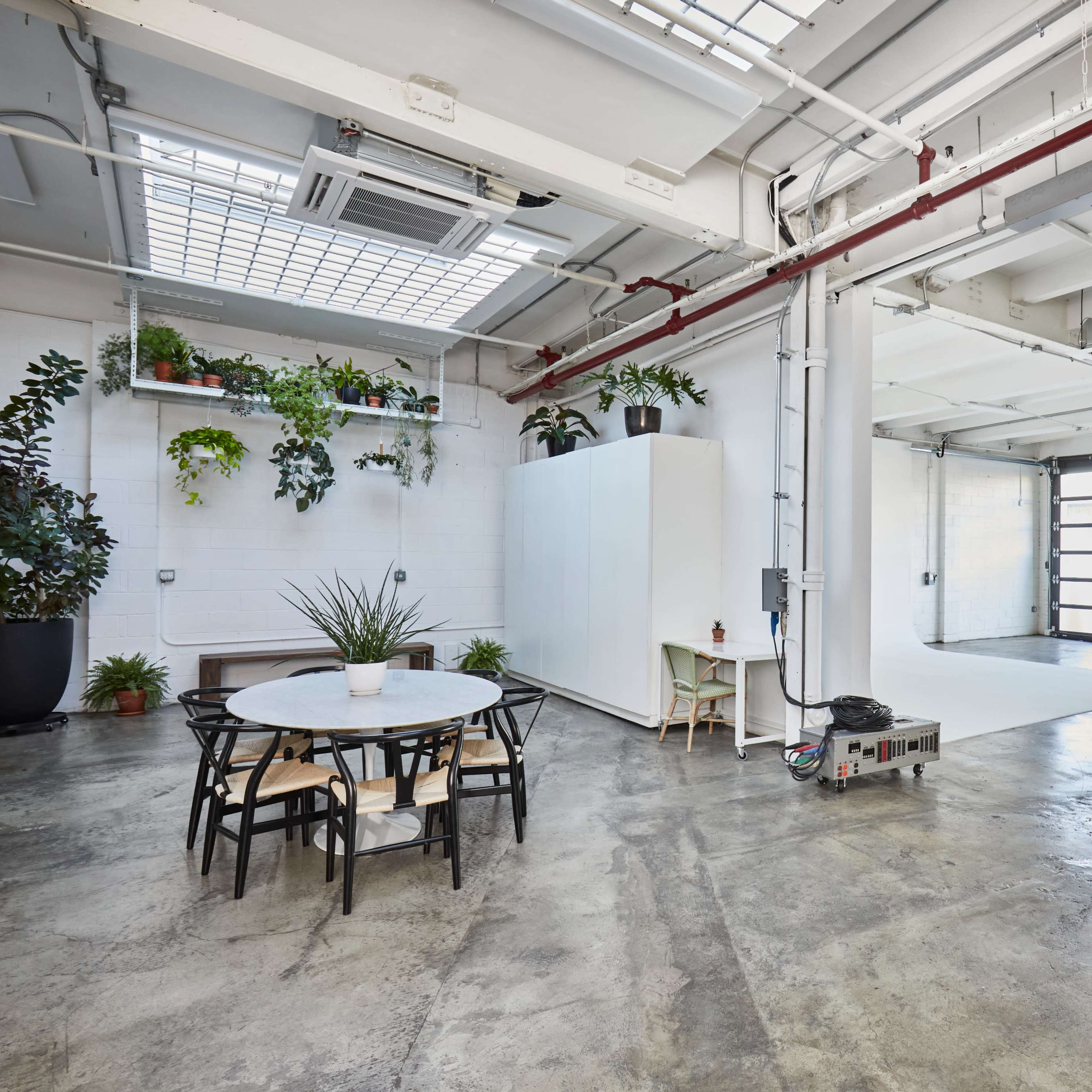 A spacious, well-lit industrial-style interior featuring a table with chairs, plants on the walls, and a large garage door opening to the outside.