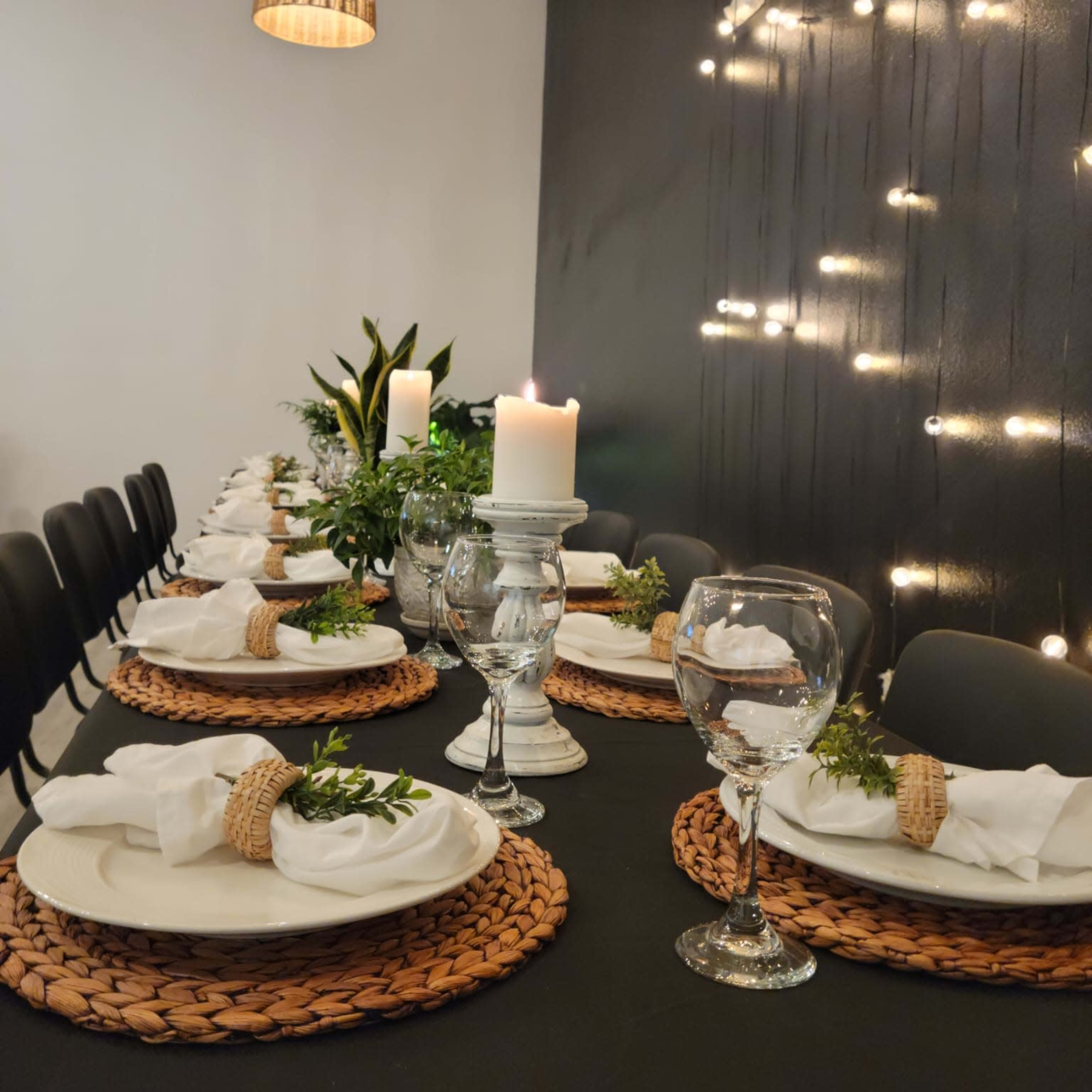 A long dining table is set with white plates, woven placemats, glasses of water, and candles, illuminated by pendant lights and string lights on a dark wall.