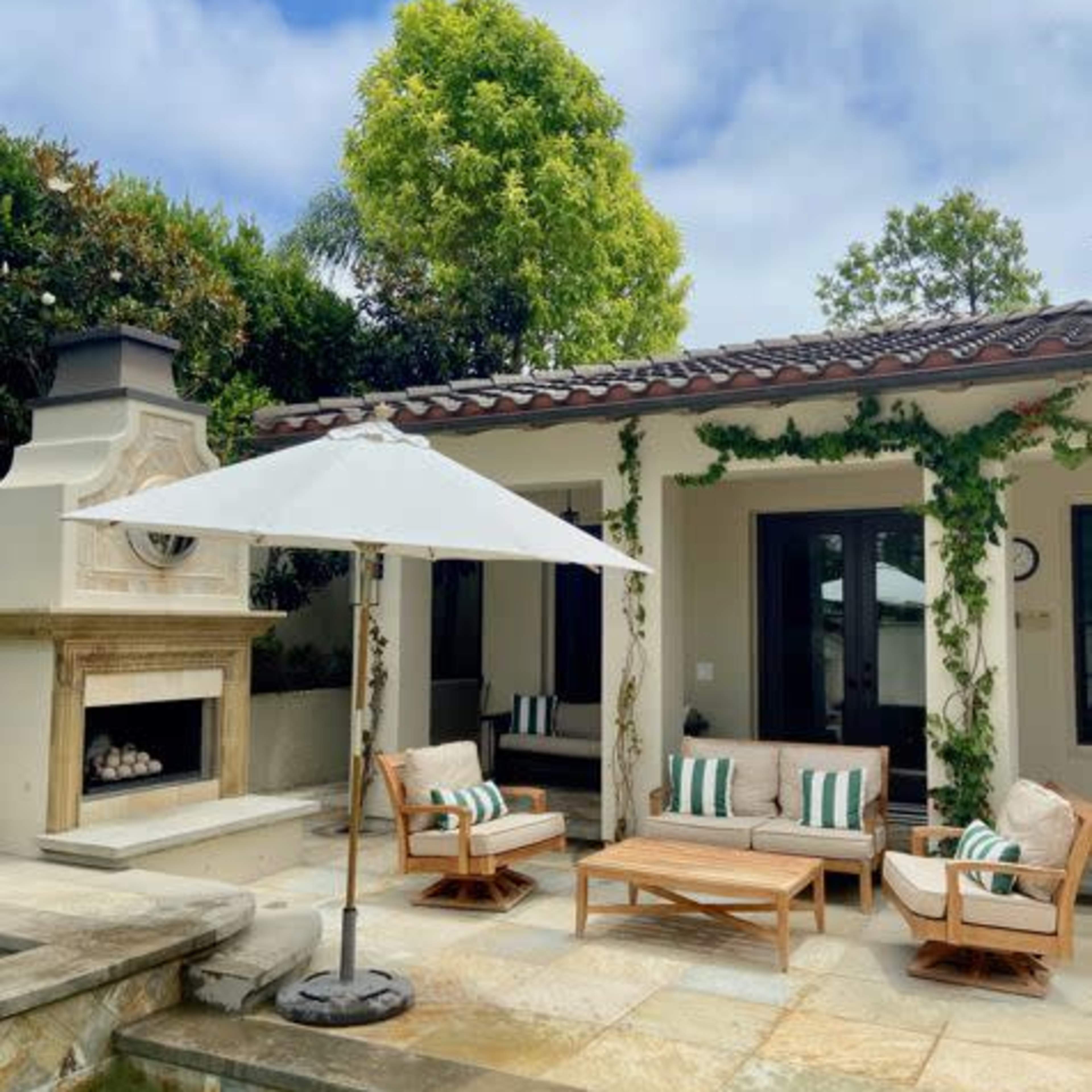 A patio area with wooden furniture, a shaded umbrella, and a stone fireplace overlooks a pool and garden.