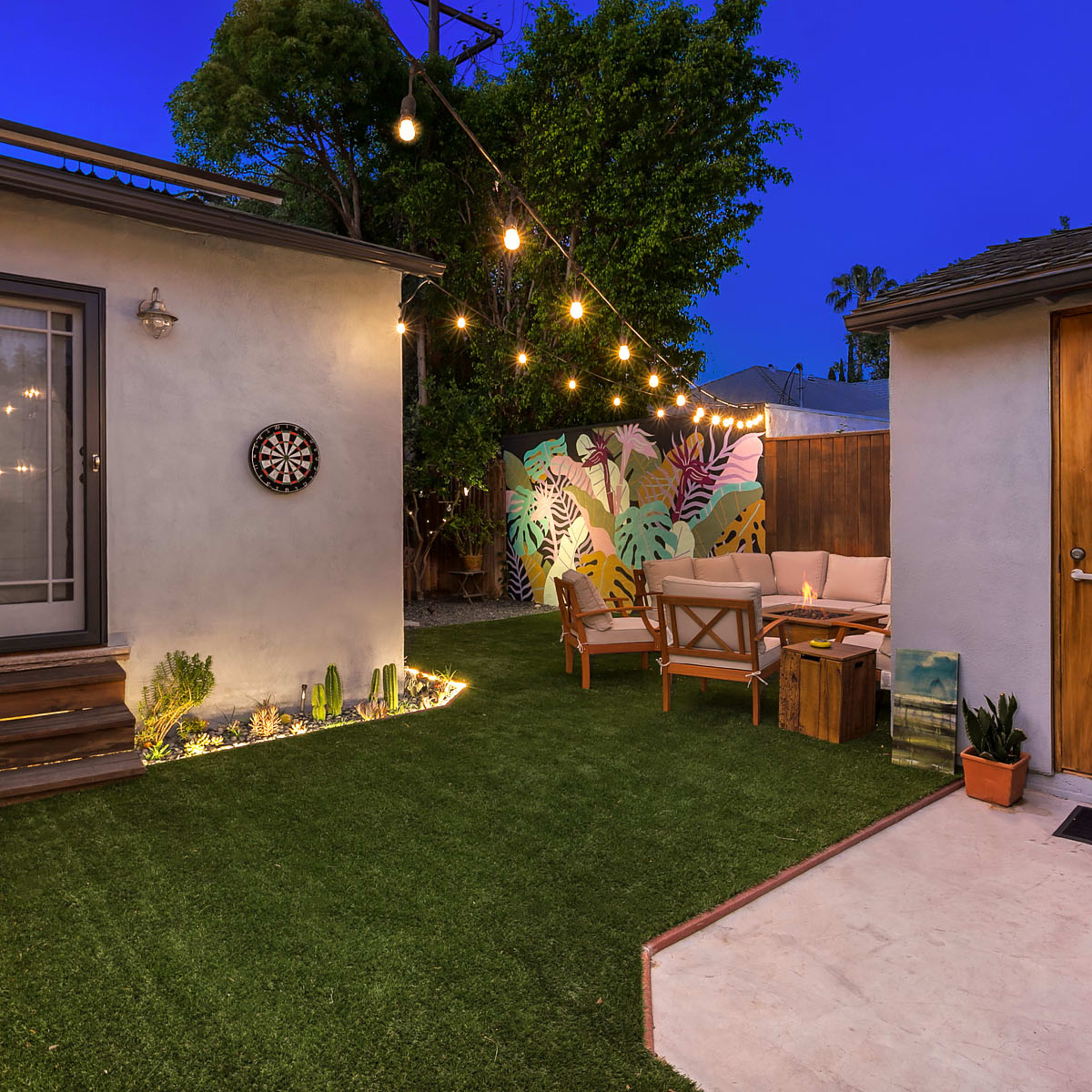 A small outdoor patio features a dartboard, string lights, artificial grass, seating area, and a colorful mural in the background.