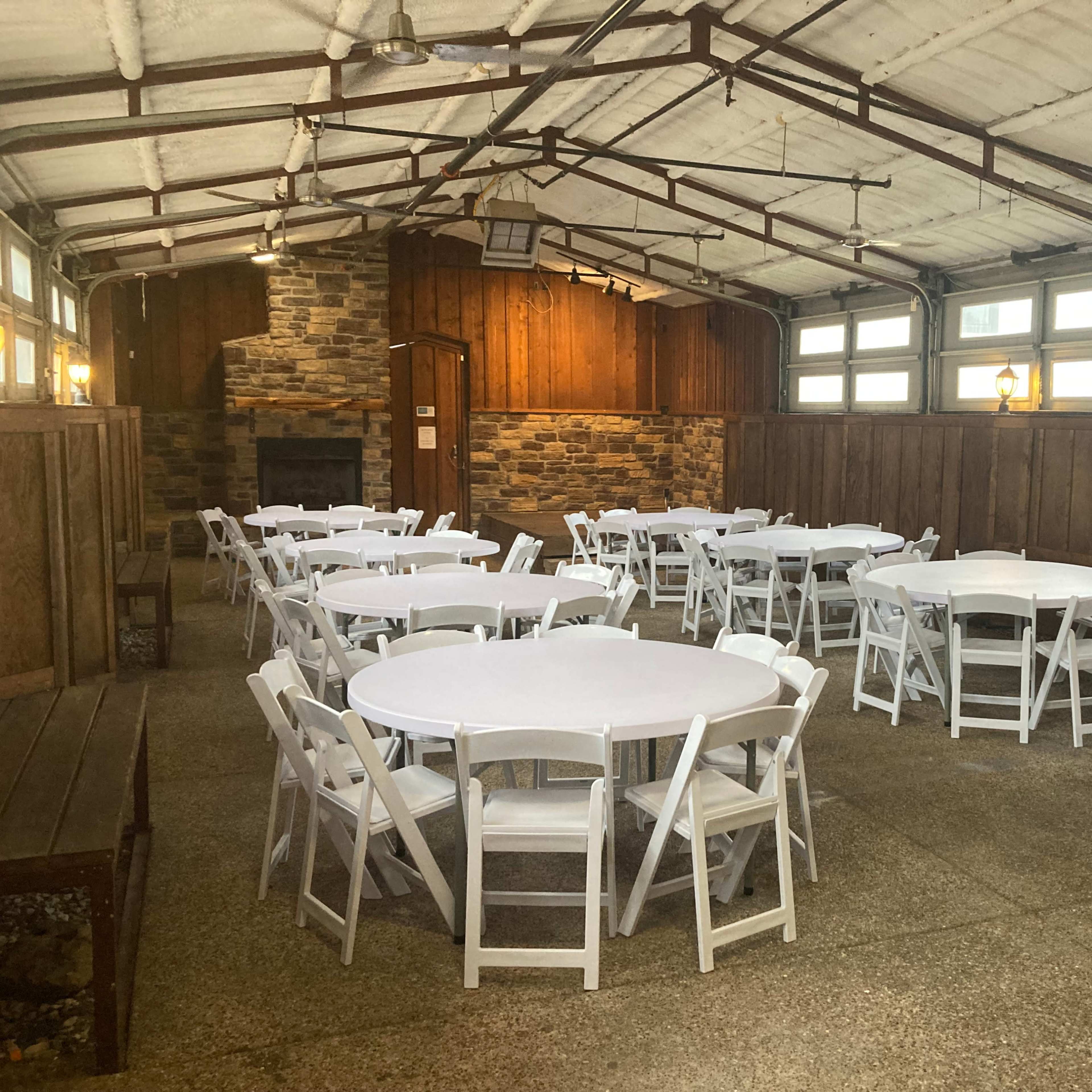A spacious indoor area with multiple round tables and white chairs arranged neatly, featuring wooden walls and a stone fireplace.