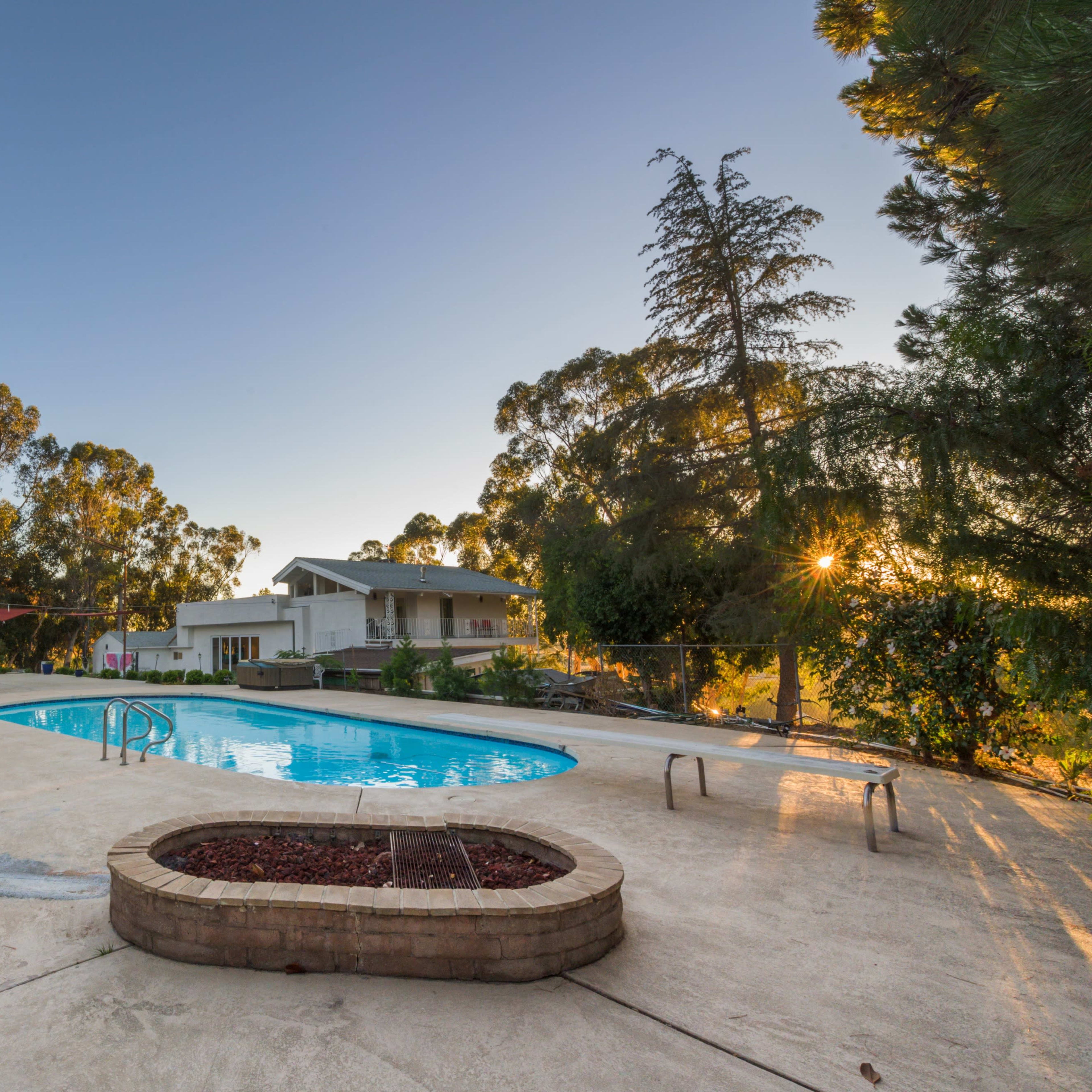 The scene shows an outdoor swimming pool surrounded by greenery and trees, with a house visible in the background.