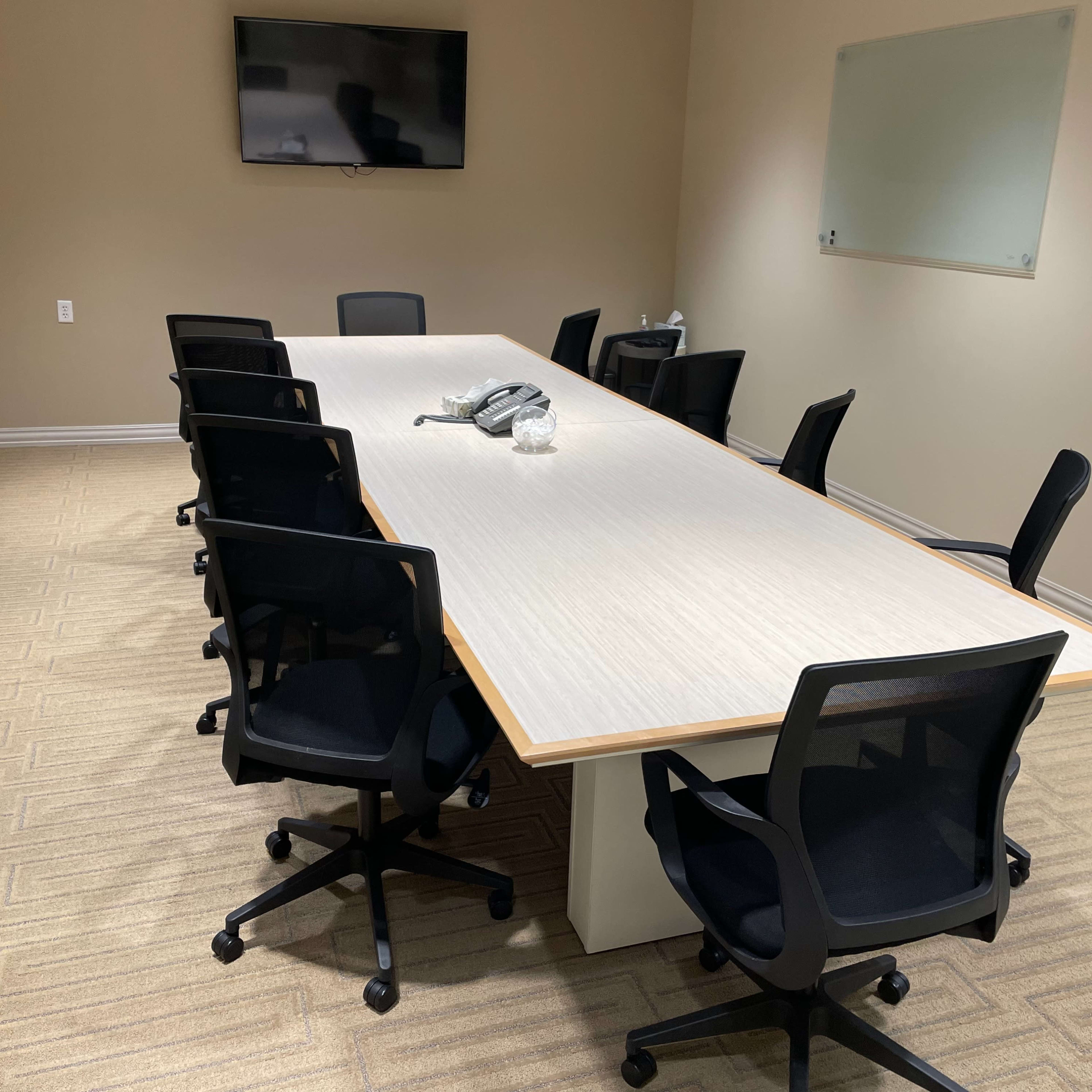 A conference room features a long table surrounded by eight black rolling chairs, with a television mounted on the wall and a whiteboard nearby.