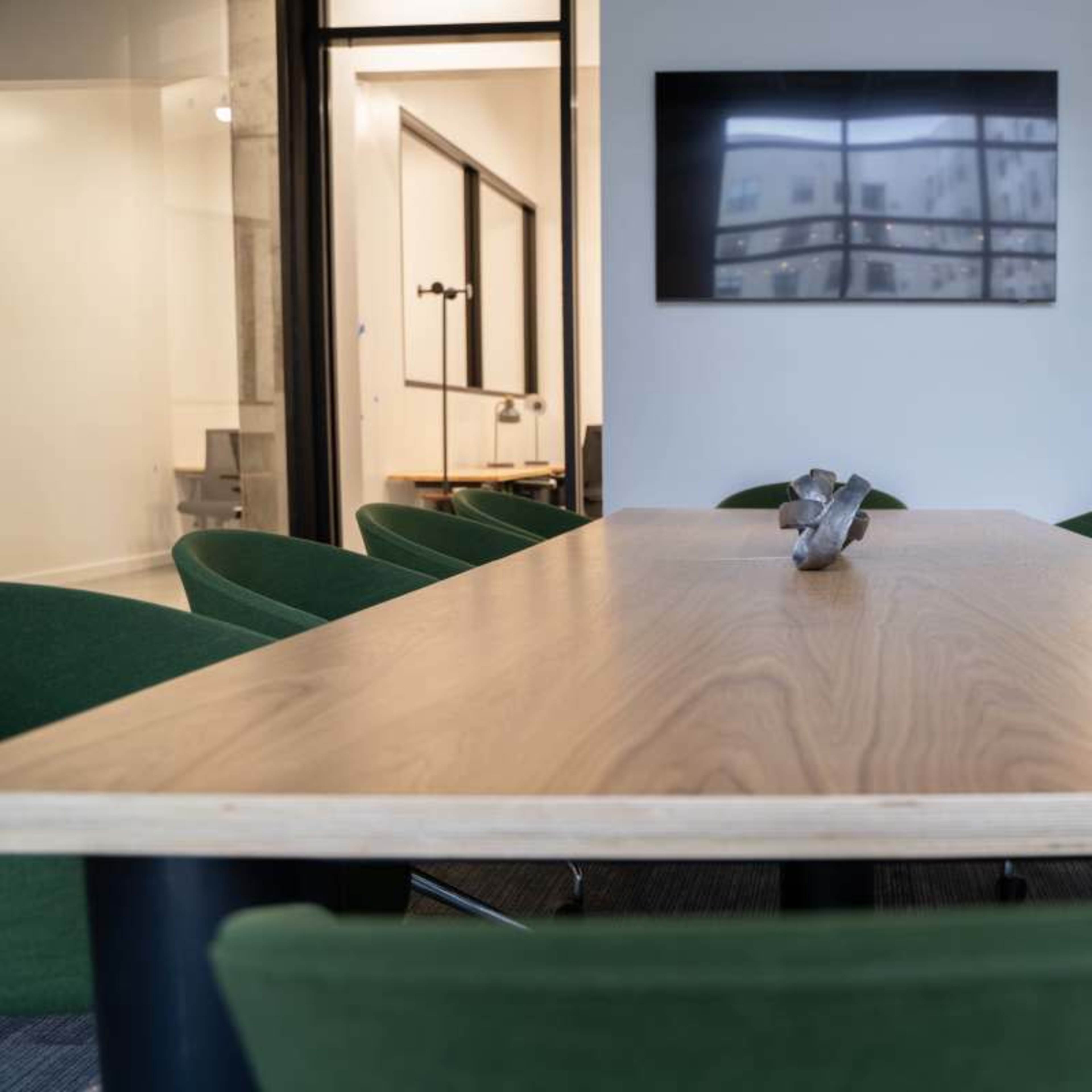 A long wooden conference table is set in a meeting room with green chairs and a large screen mounted on the wall.