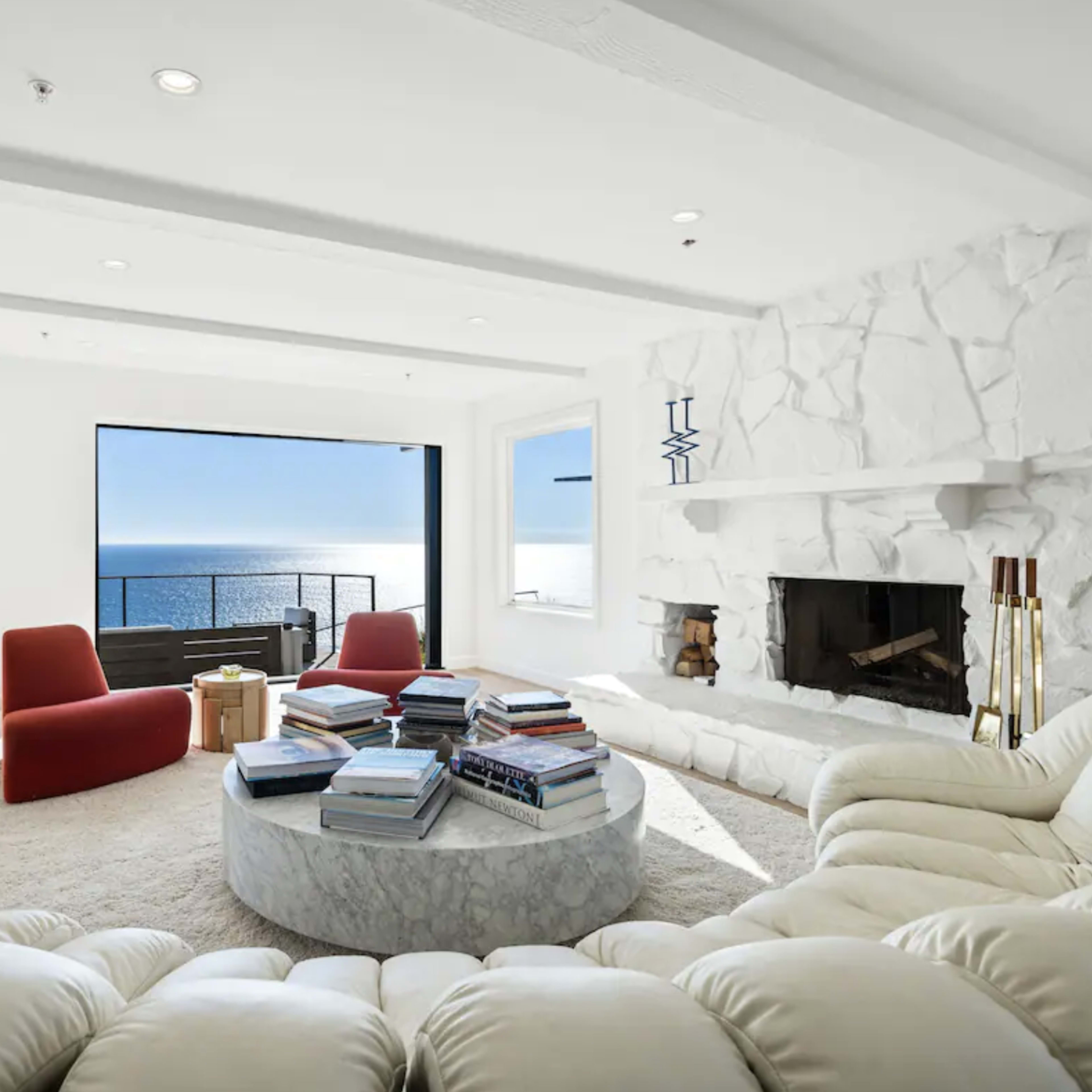 The room features a circular marble coffee table surrounded by a white sectional sofa and red chairs, with large windows showcasing an ocean view.