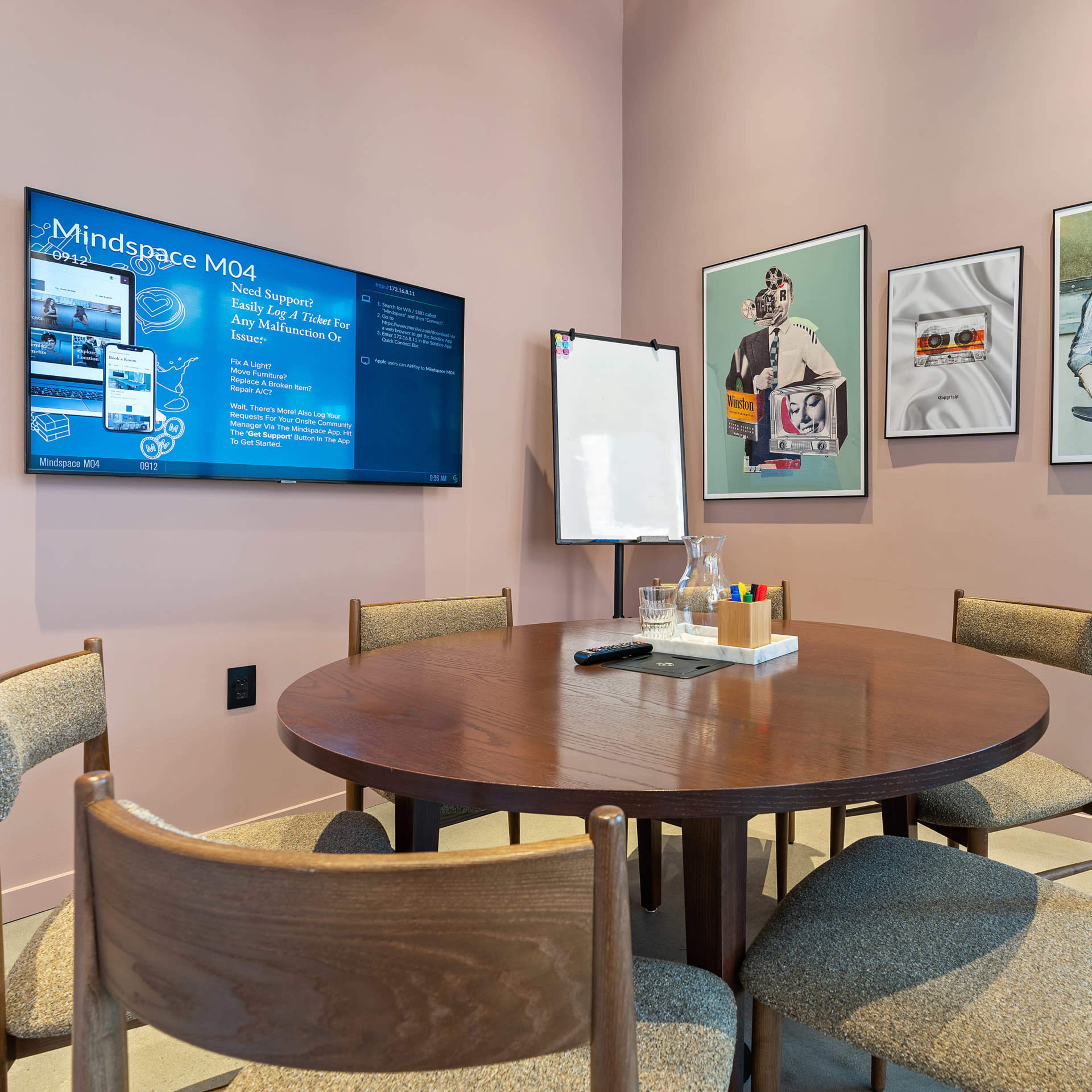 A modern meeting room with a round wooden table, four chairs, a digital screen displaying information, a whiteboard, art on the walls, and a potted plant in the corner.