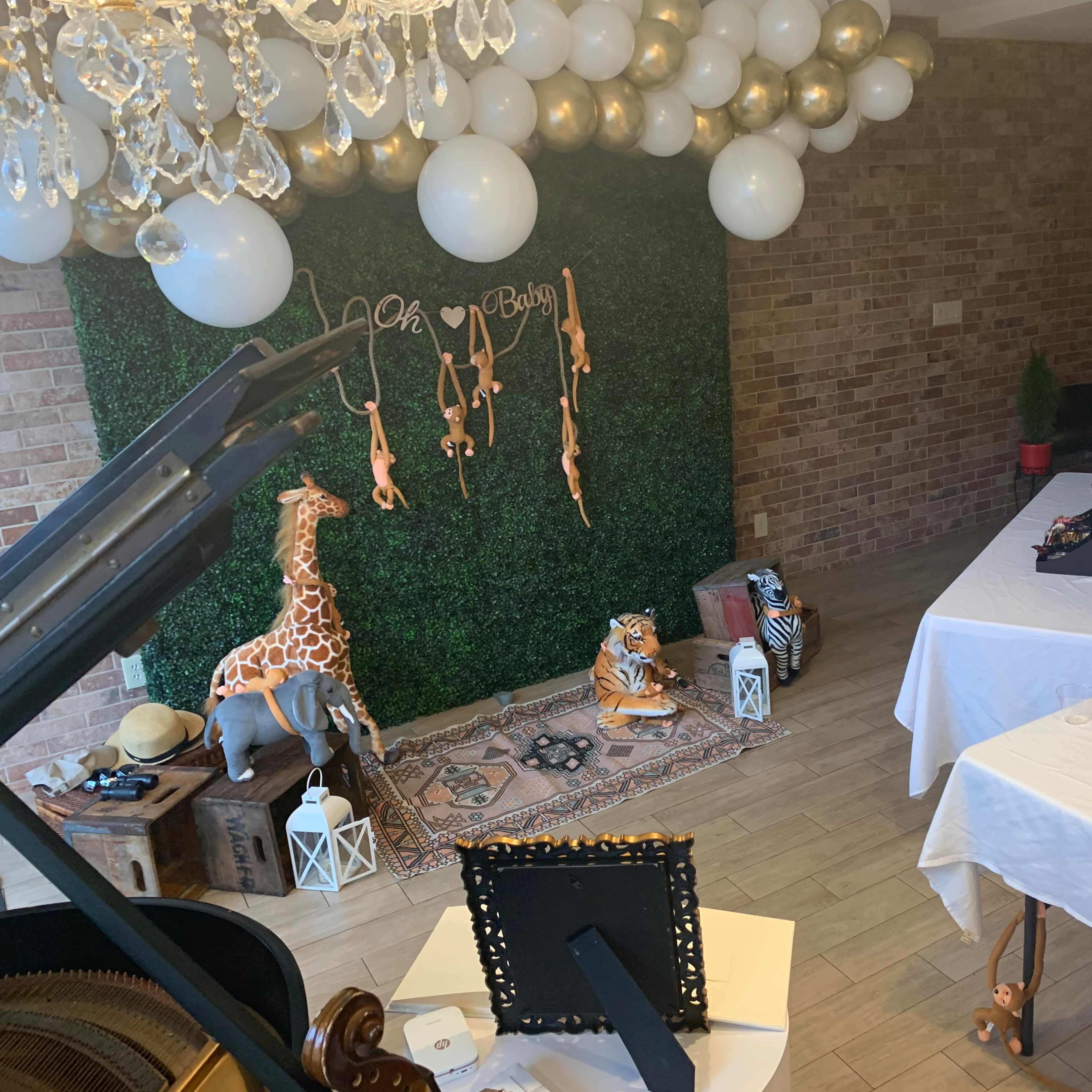 The scene features a decorated space with a greenery wall, animal-themed decorations, a piano, and tables set with food and drinks for an event.