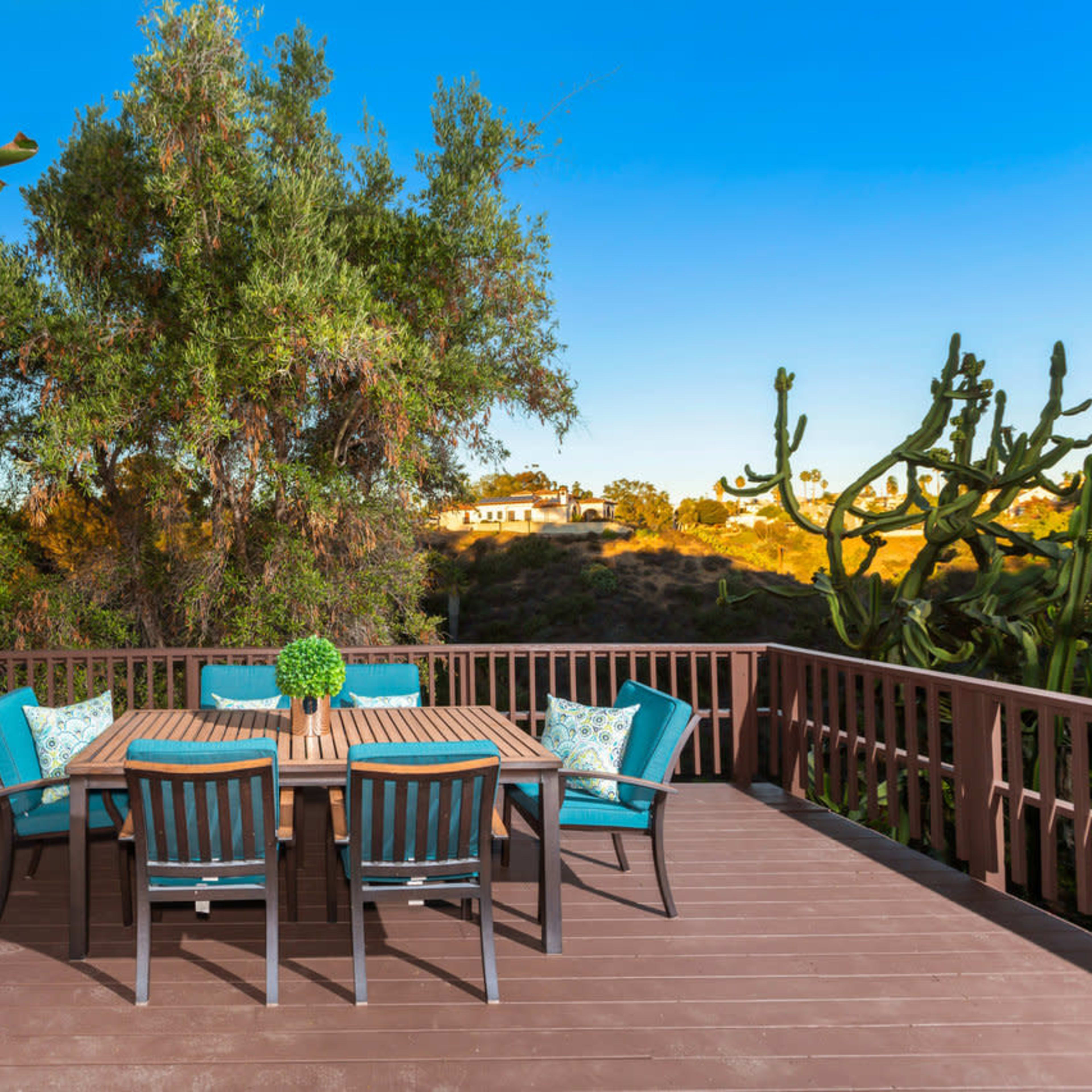 A wooden deck with a dining table and six chairs is surrounded by greenery and cacti in a residential area.