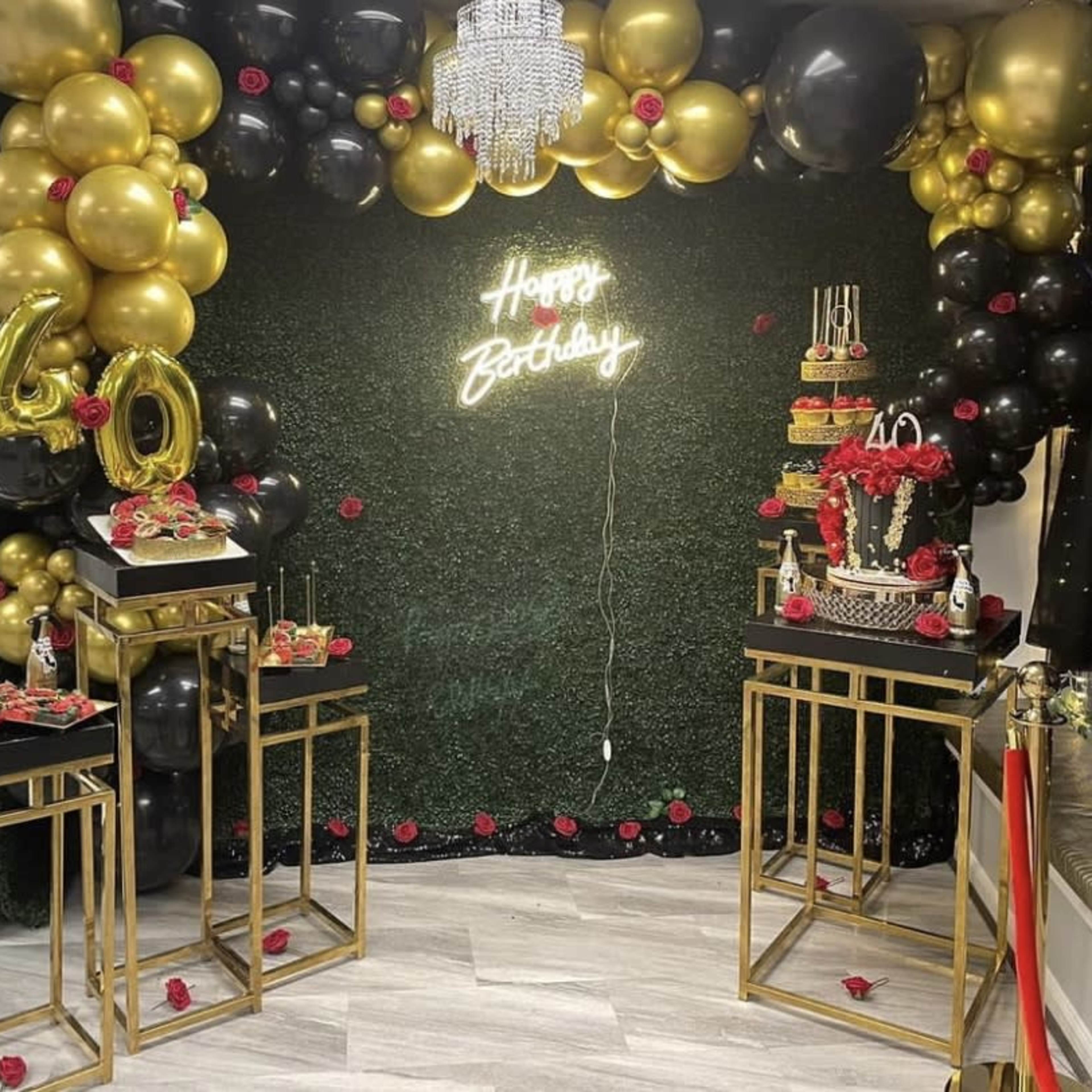 A decorated birthday party setup featuring a neon sign that says "Happy Birthday," a large cake on a stand, and a backdrop of black, gold, and red balloons.