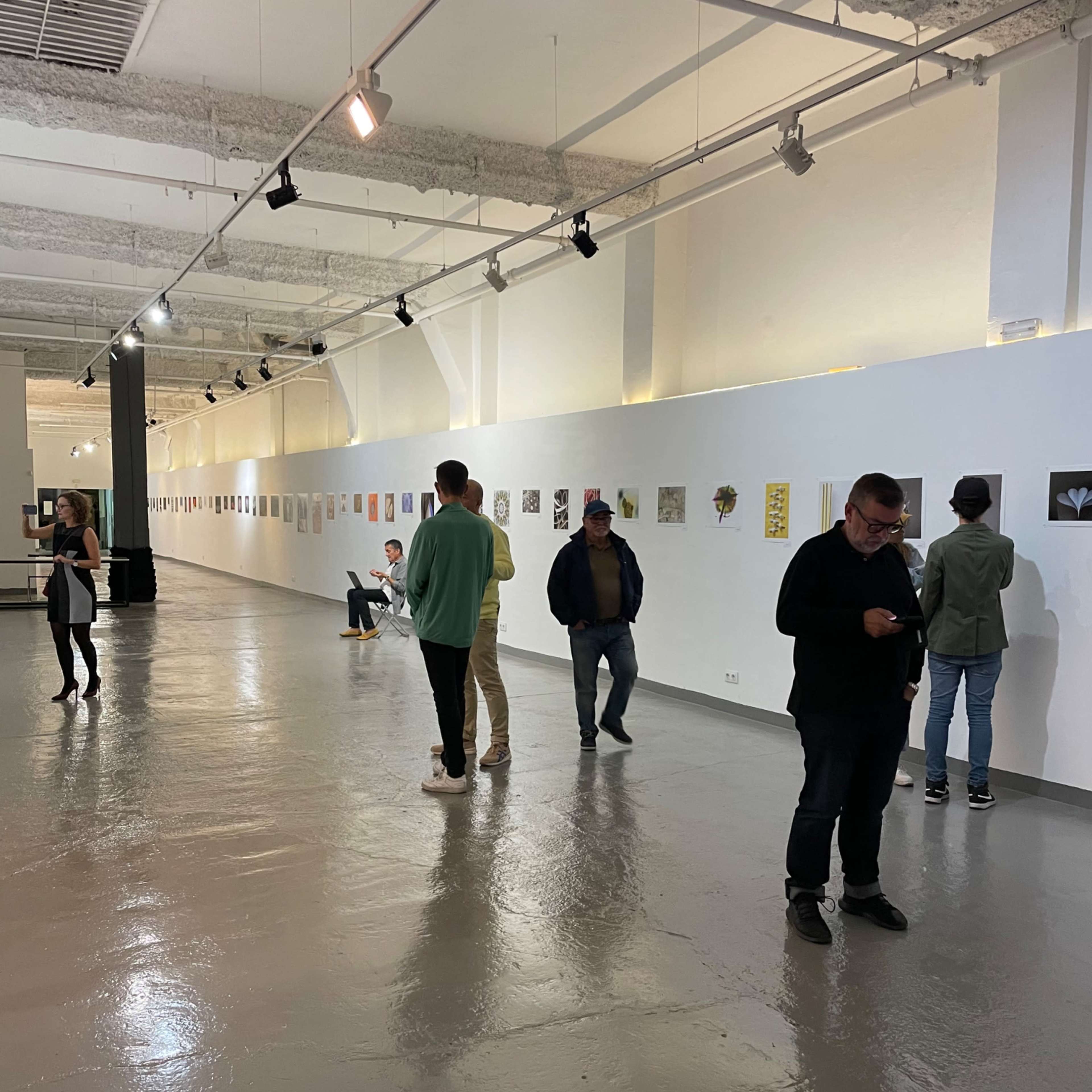An art gallery with several people examining artworks displayed on the walls in a spacious, well-lit area.