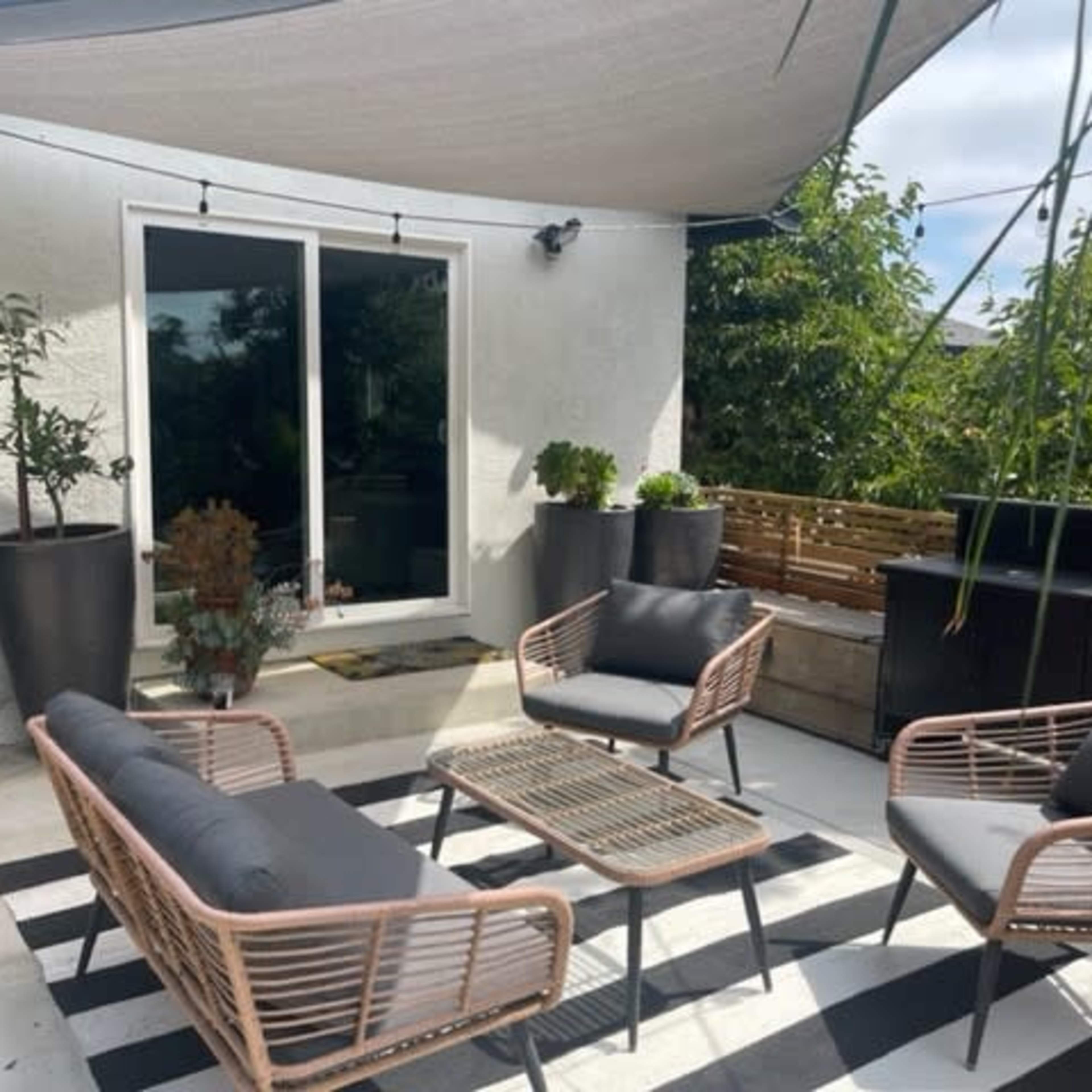 A patio area features a table and four chairs arranged on a black and white striped rug, surrounded by potted plants and a grill.