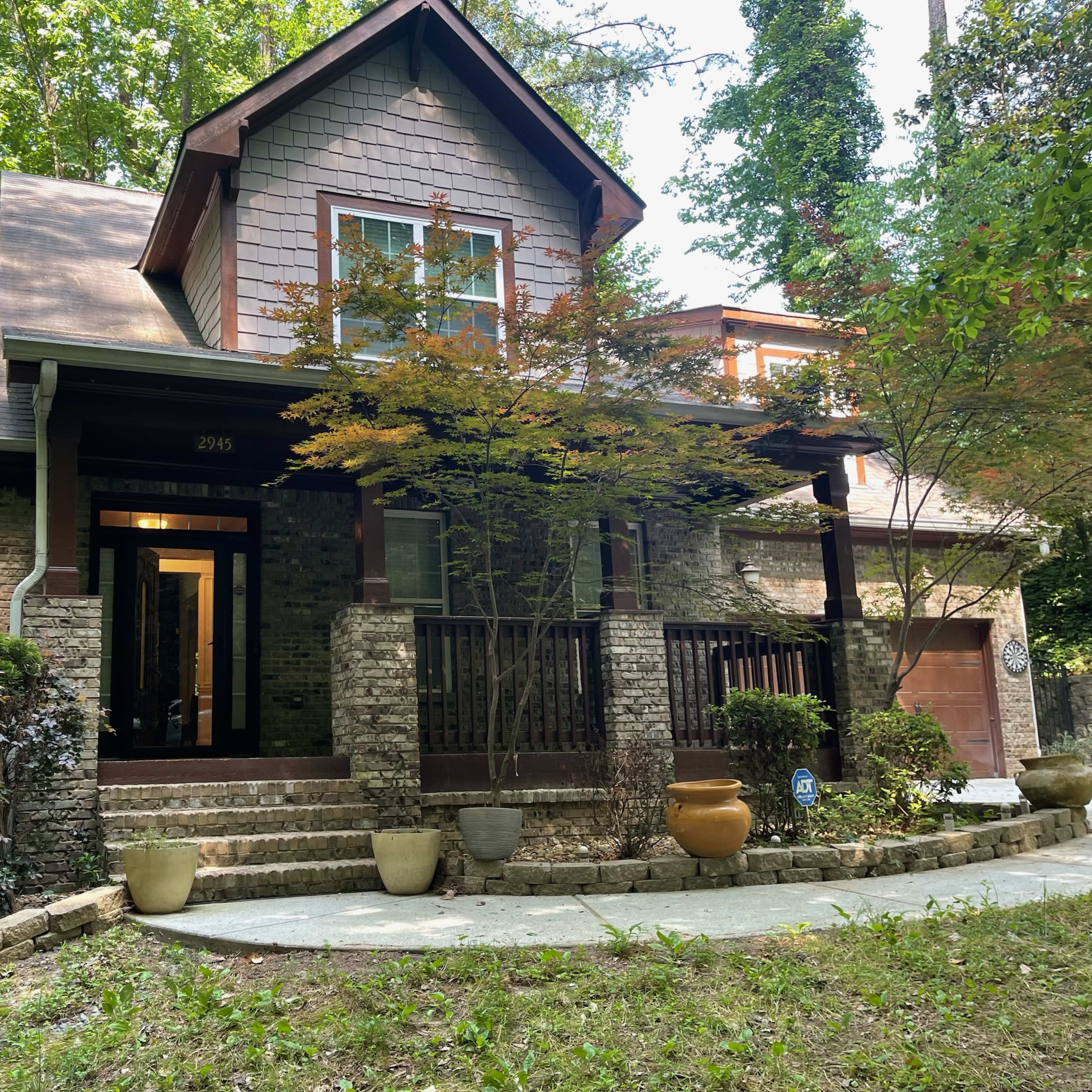 A two-story house with a stone facade and a covered porch is surrounded by greenery and has multiple planters at the entrance.
