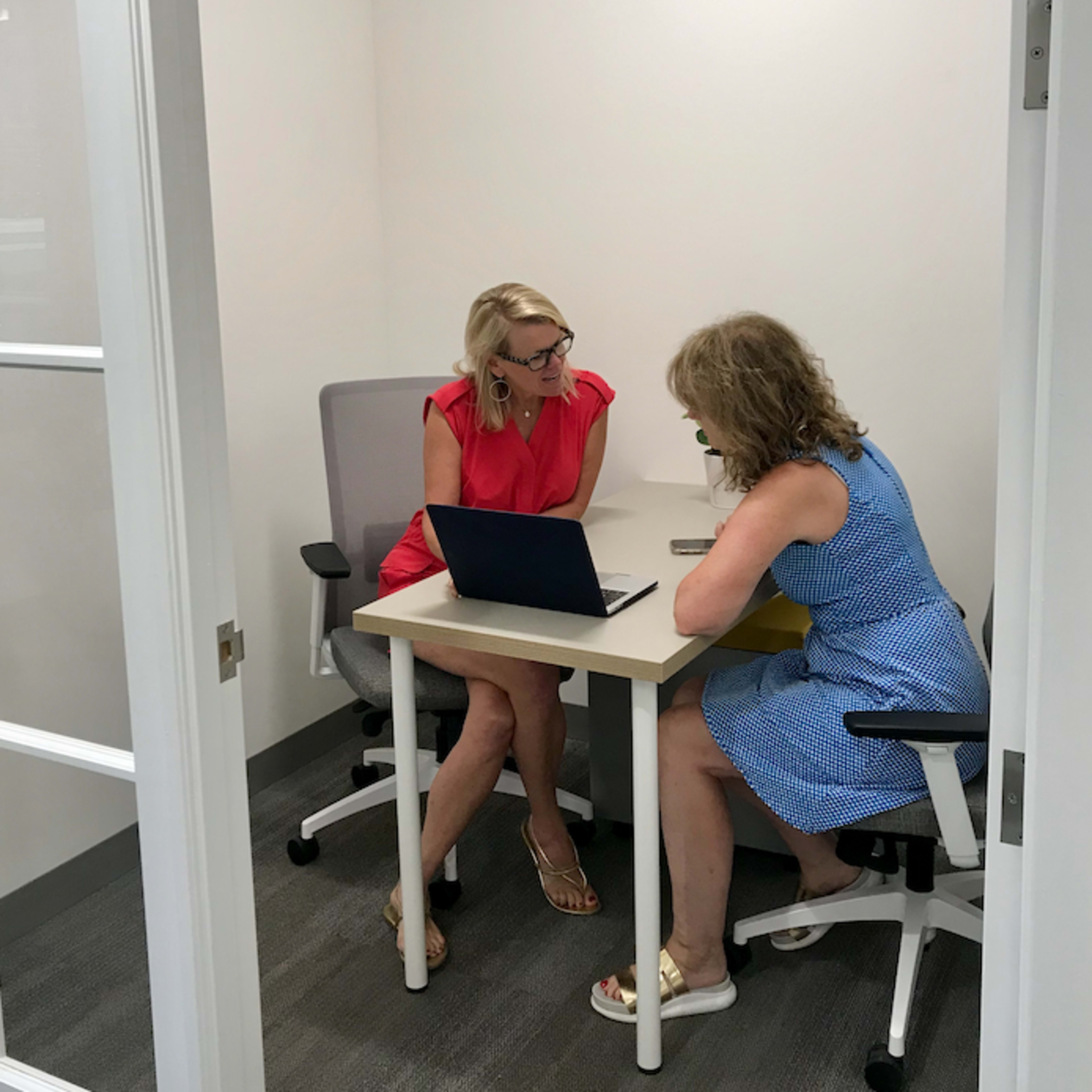 Two women are seated at a small table in an office meeting room, engaged in discussion while one uses a laptop.