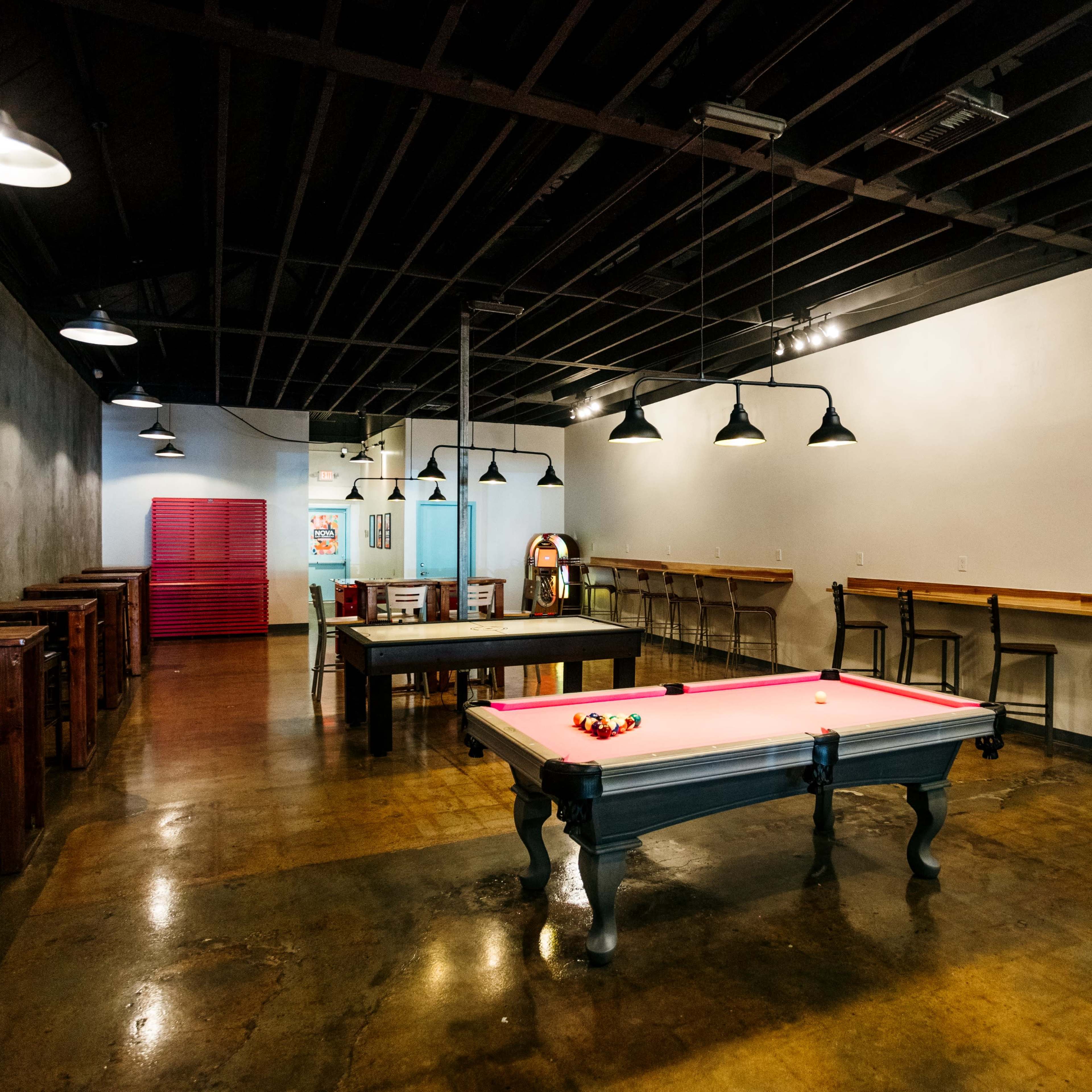 A spacious game room features a pool table, a jukebox, and several tables with chairs arranged around a polished concrete floor.