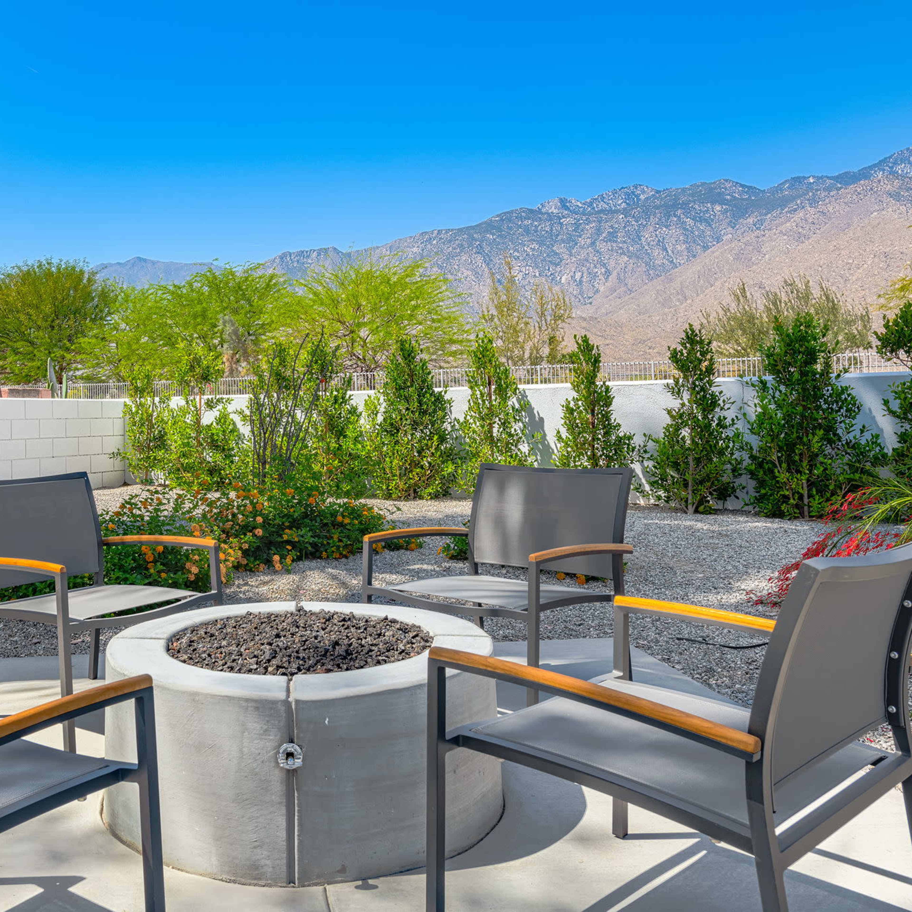 A circular fire pit surrounded by four gray chairs sits in a landscaped backyard with mountains in the background.