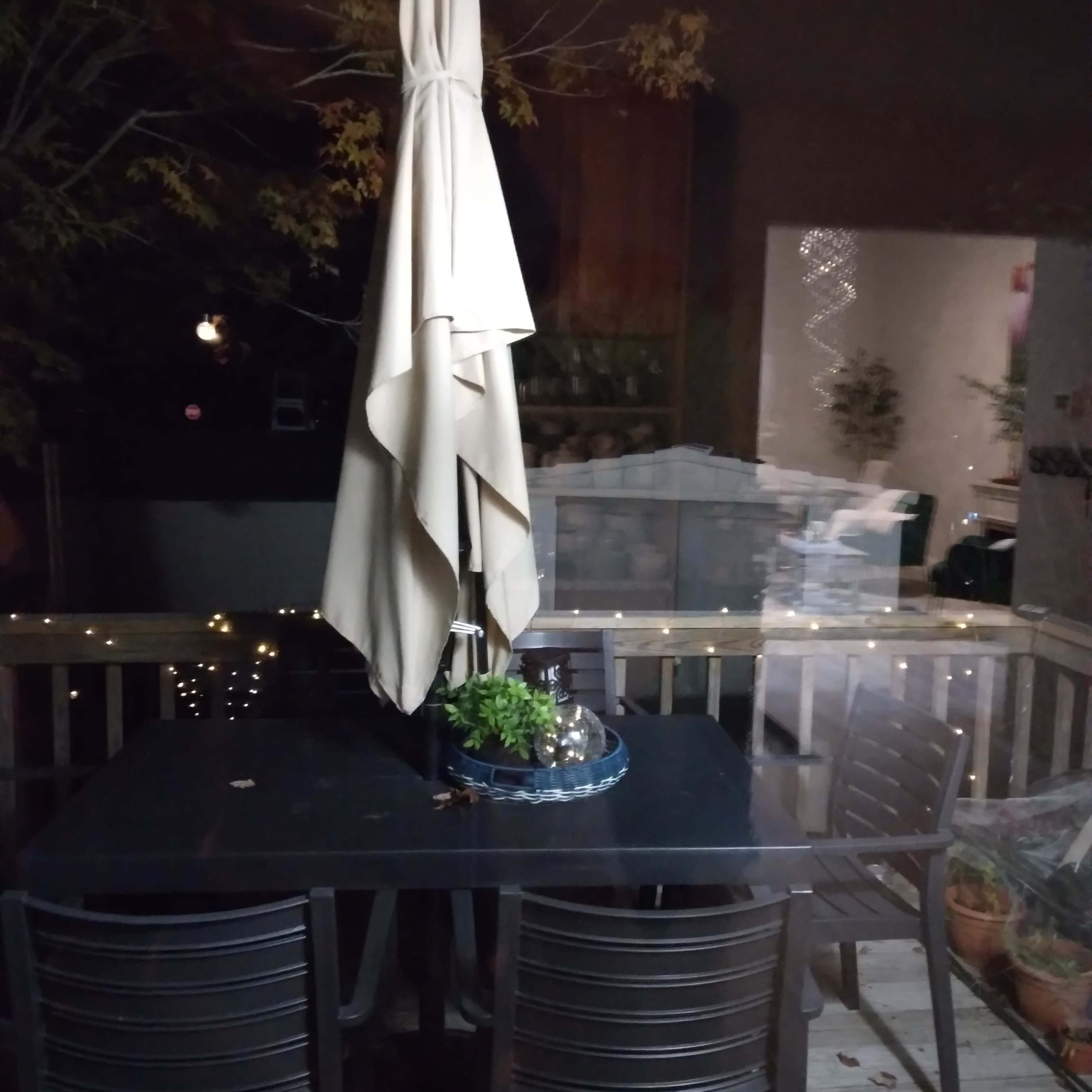 A patio table with a beige umbrella stands surrounded by four wooden chairs on a deck at night.