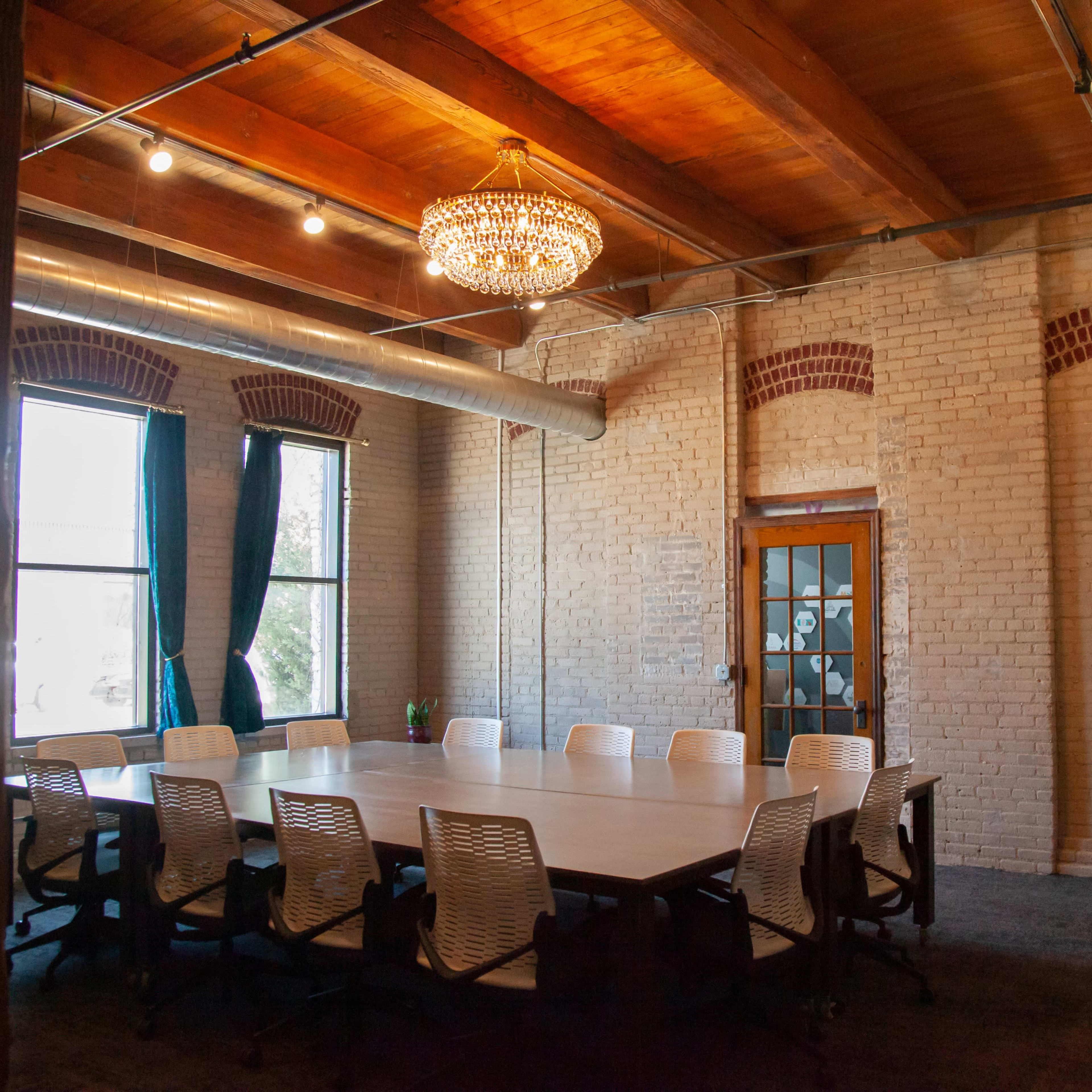 A conference room features a long table surrounded by chairs, with large windows, exposed brick walls, and a chandelier overhead.