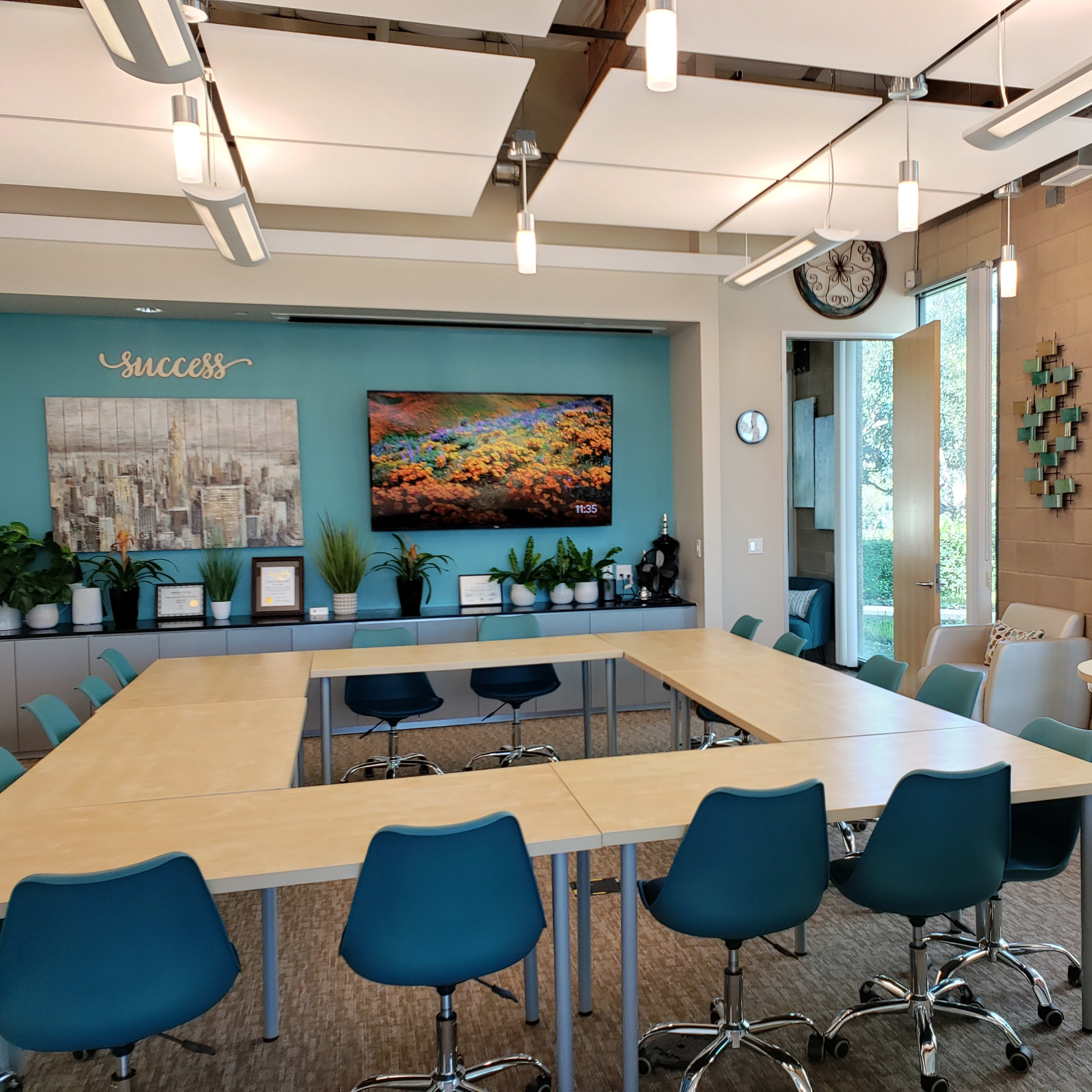 A modern conference room with multiple tables arranged in a U-shape, surrounded by teal chairs, and features large windows with plants and artwork on the walls.
