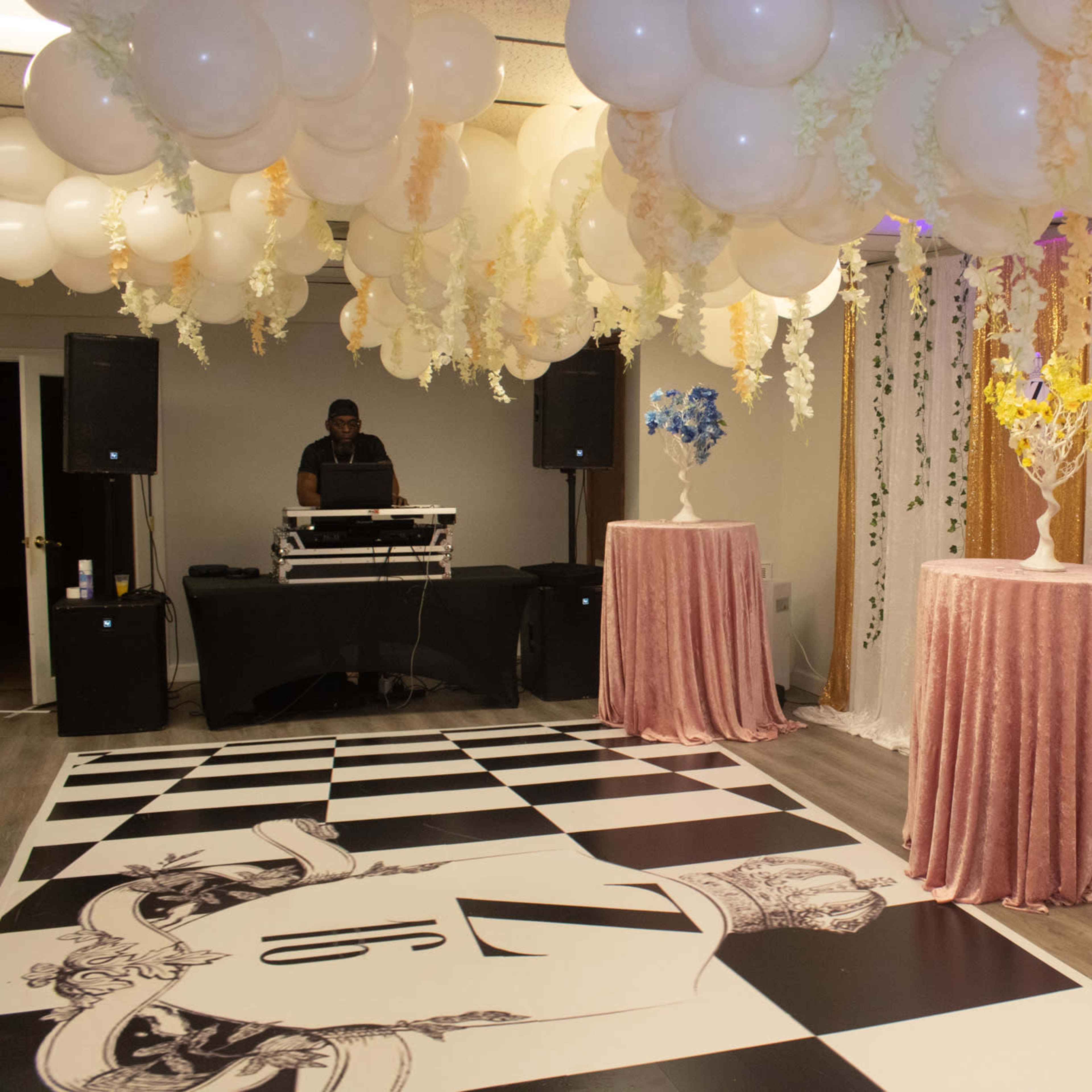 The room features a checkered dance floor with two circular tables adorned with floral arrangements and a DJ setup in the background, all beneath a canopy of balloons and draping.