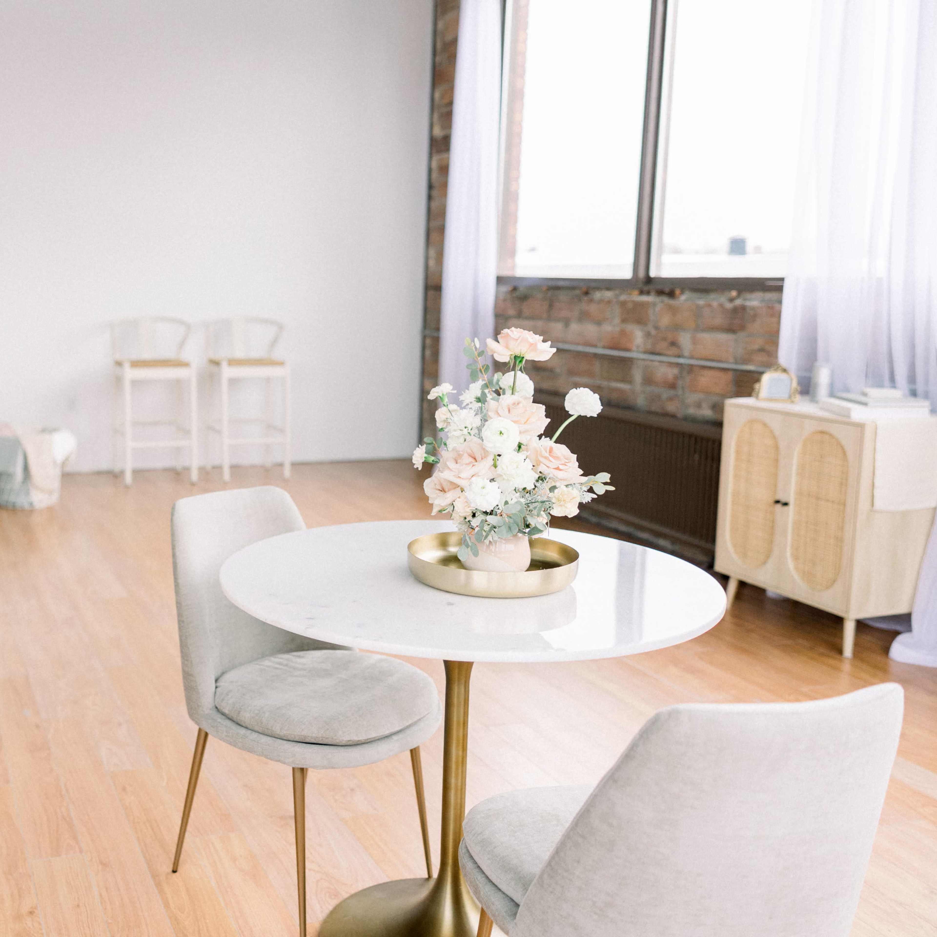 A modern, minimalist interior with a round table and two chairs, featuring a floral centerpiece, along with a sofa and two additional chairs in the background.