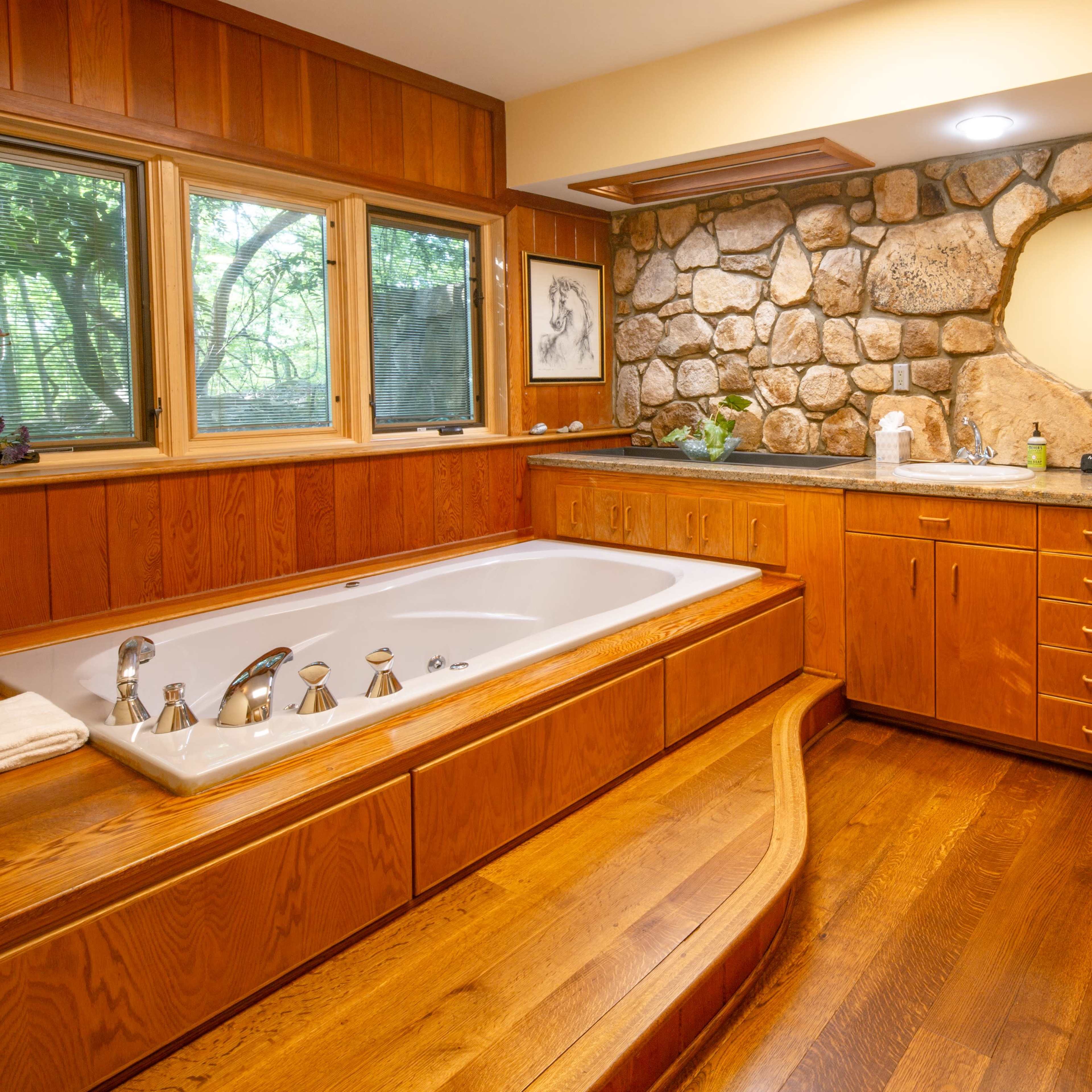 A bathroom with a large stone wall, a white bathtub, wooden cabinetry, and double sinks under bright lighting.