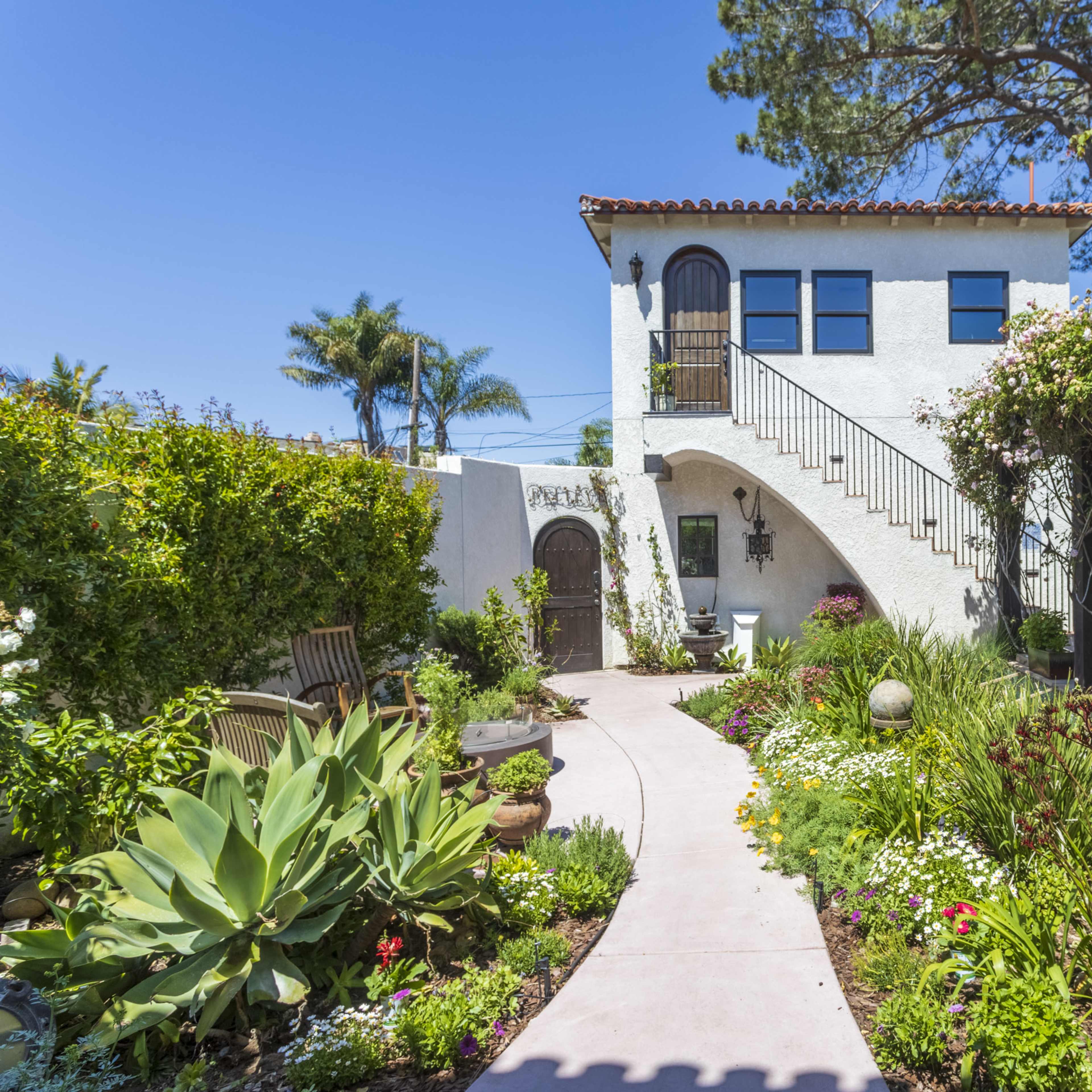 A pathway leads through a garden filled with various plants and flowers to a two-story white house with a staircase and a covered seating area adorned with greenery.