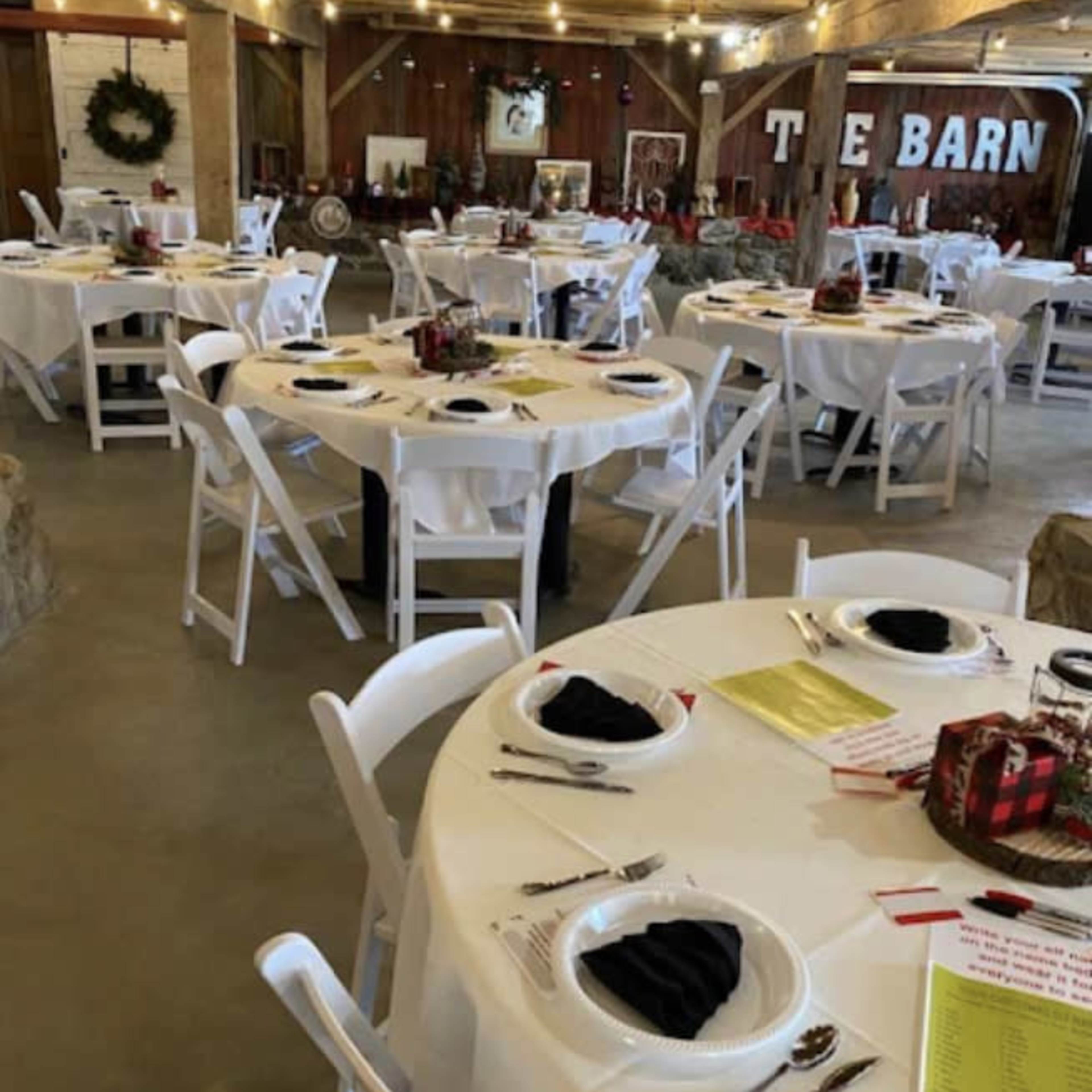 A decorated barn interior with several round tables set for a meal, each featuring white tablecloths and black napkins.