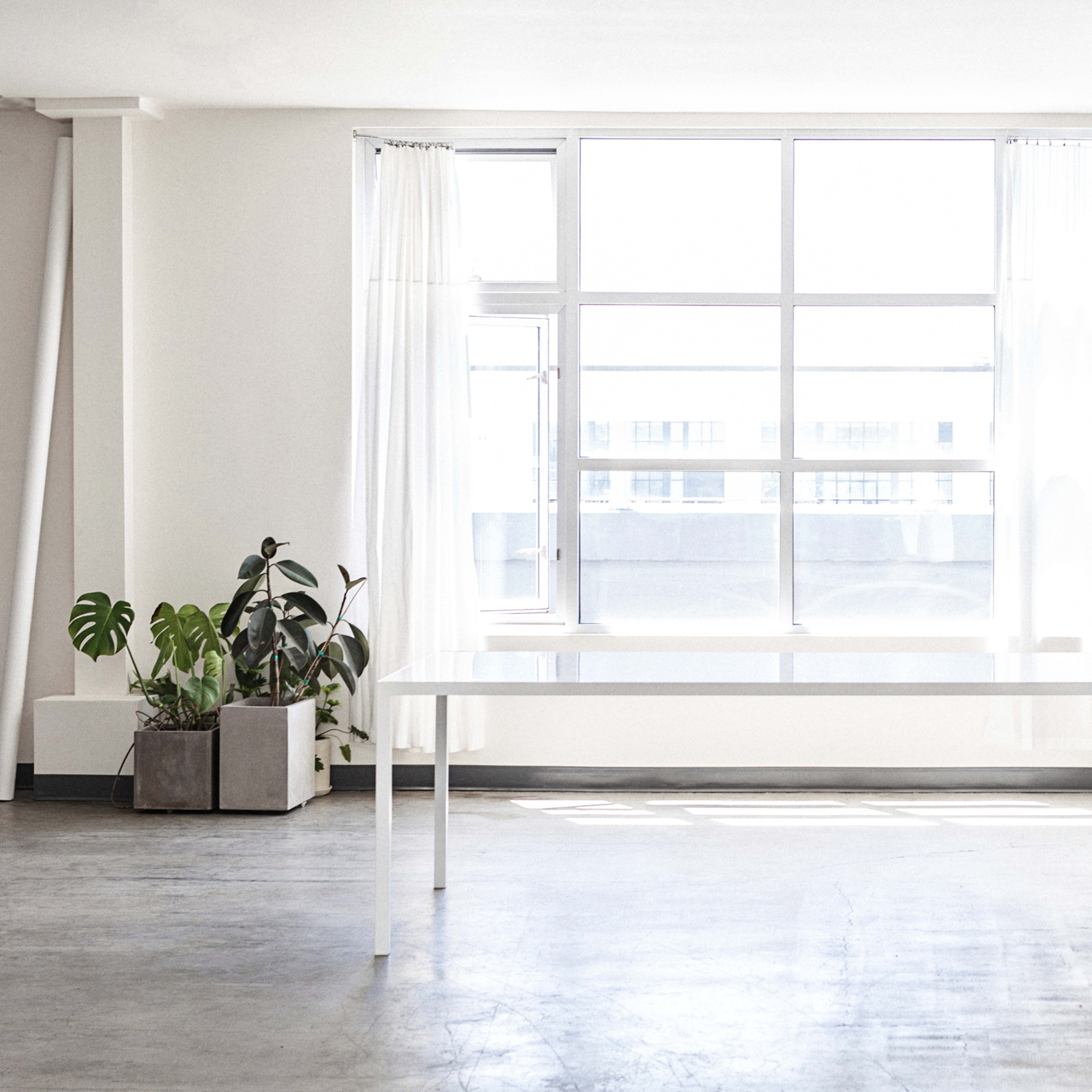 A bright, minimalistic room features a large window, a white table, a single chair, and a potted plant.