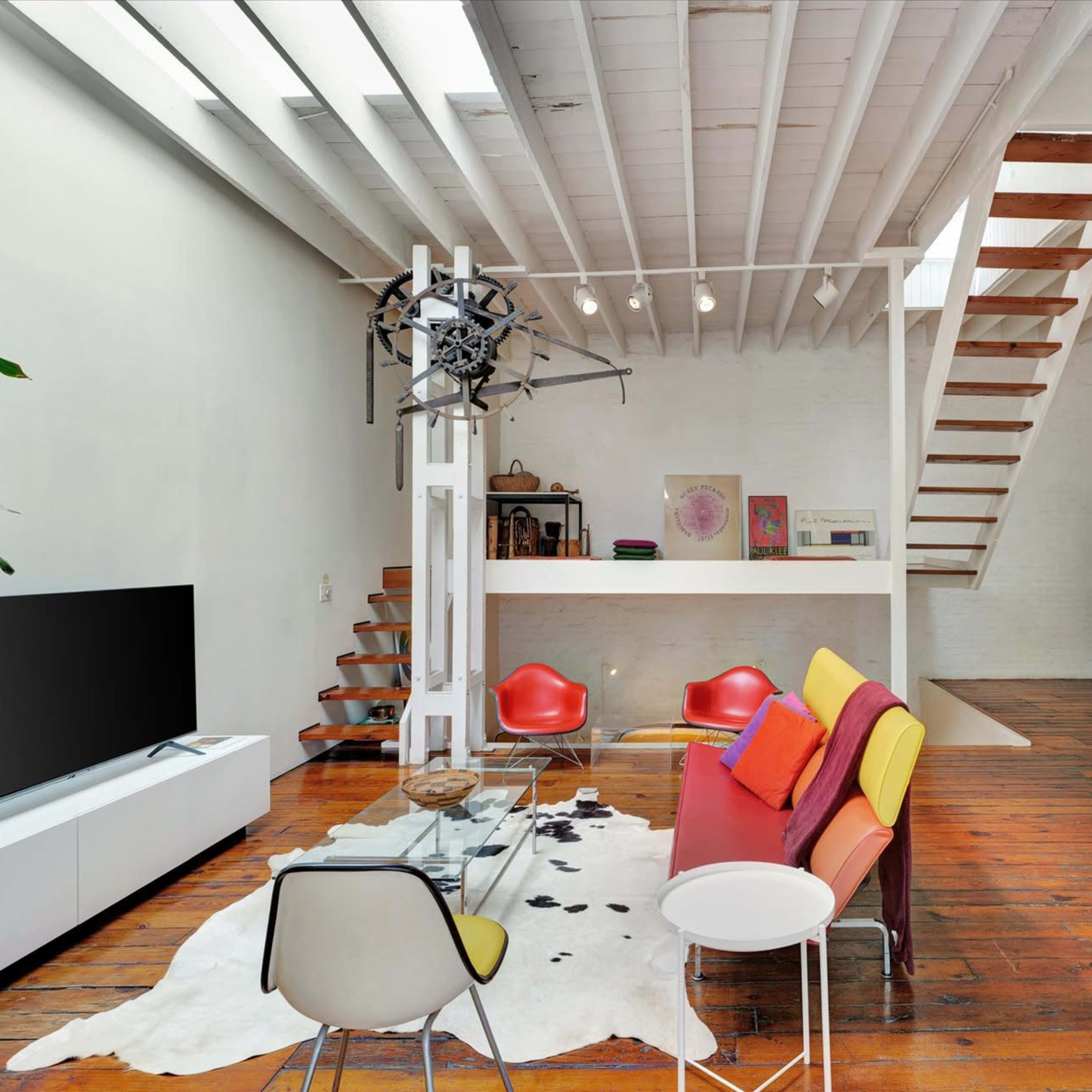 A modern living space with wooden floors, a staircase, a glass coffee table, and colorful seating arranged around a cowhide rug.