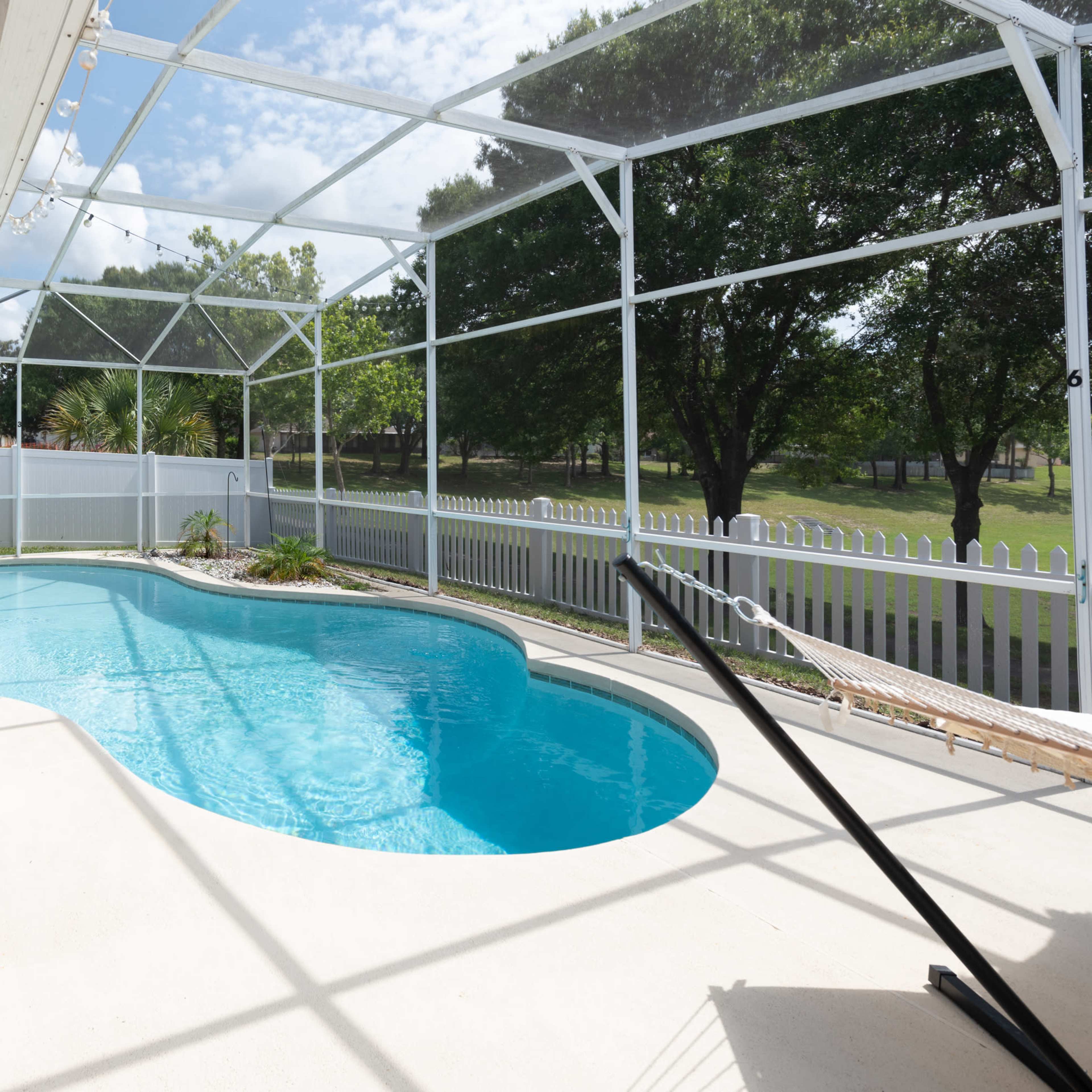 A screened-in patio area with a swimming pool surrounded by greenery and a white fence.