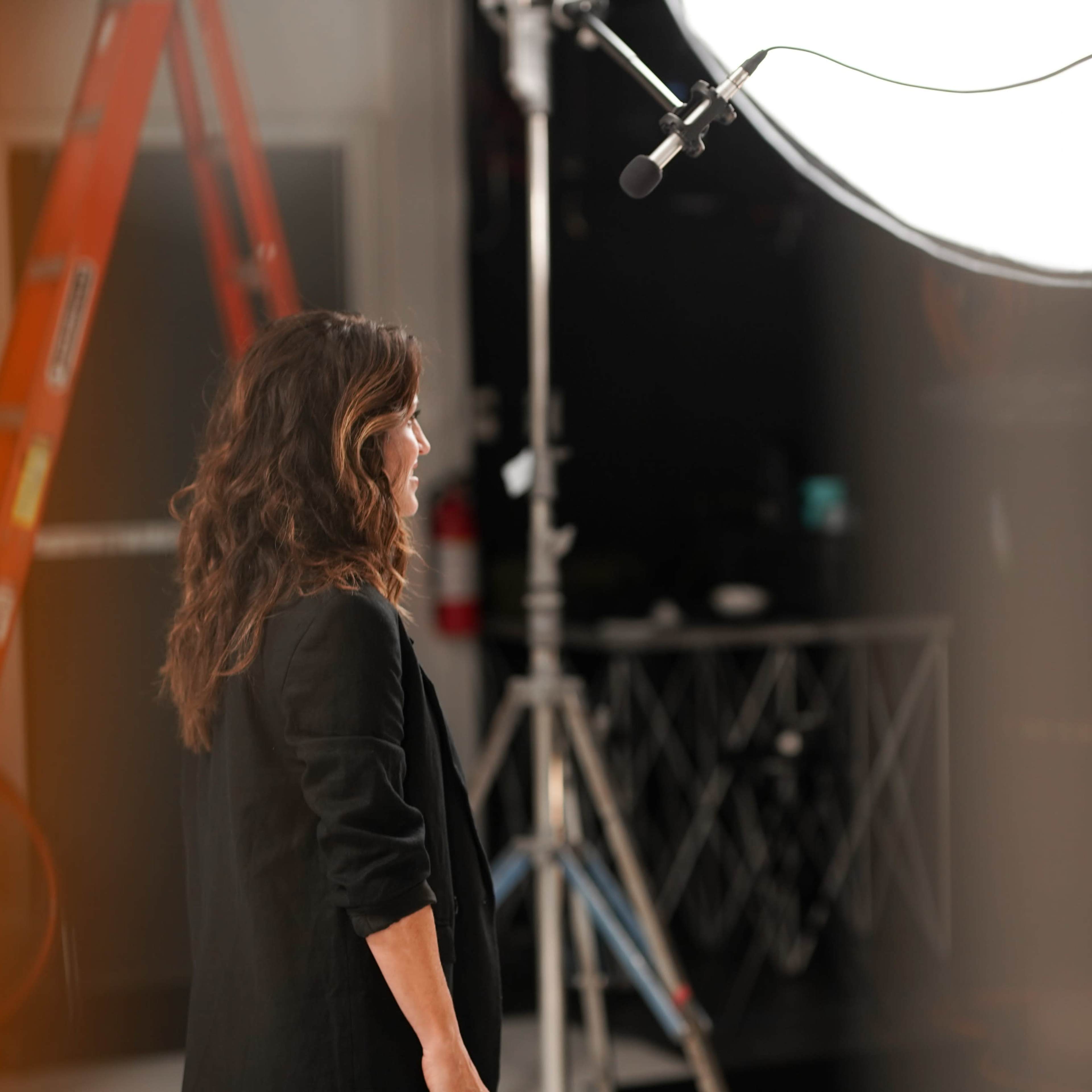 A woman stands near a large light source in a studio, with a ladder and studio equipment visible in the background.
