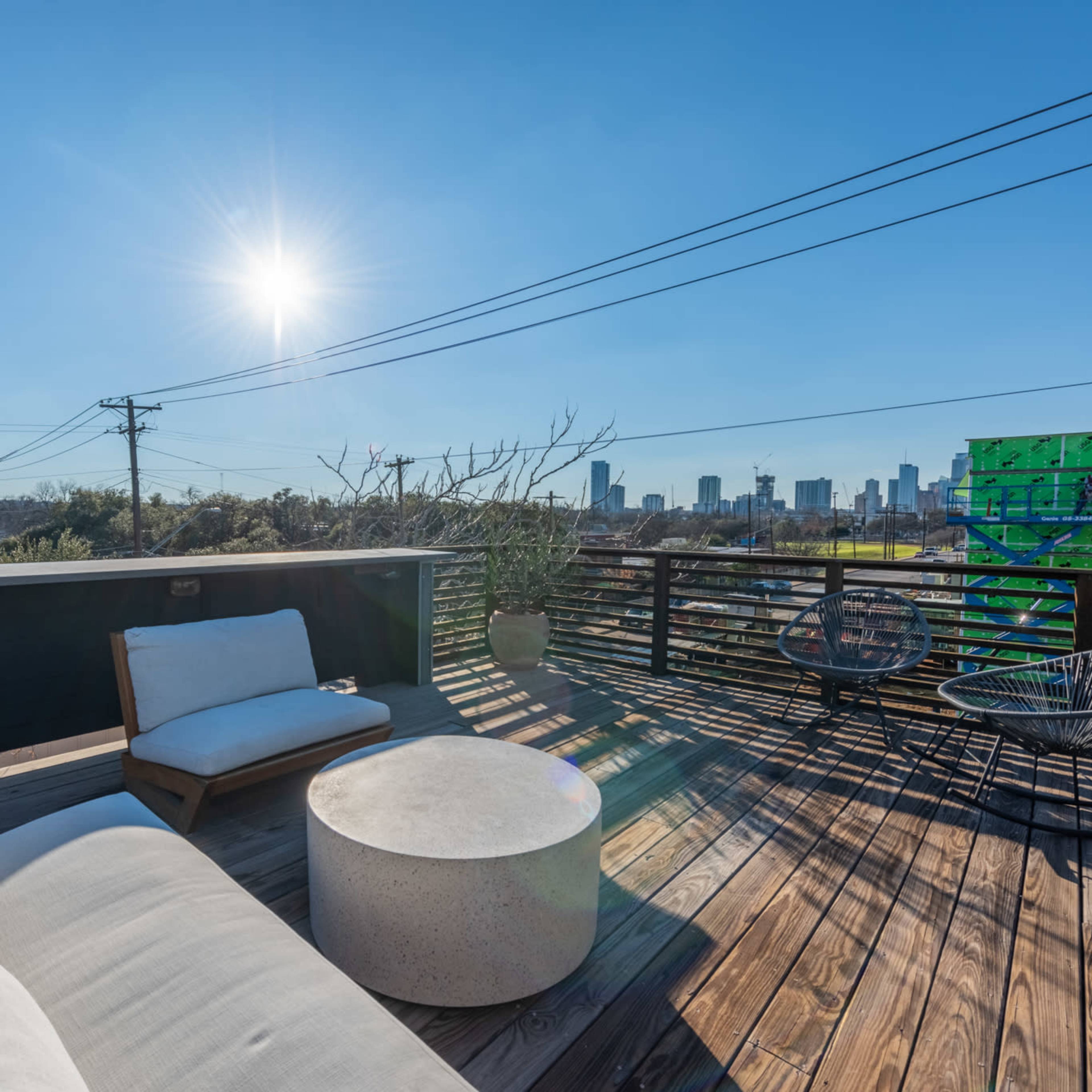 A rooftop terrace features a seating area with a concrete table and views of a city skyline under a clear blue sky.